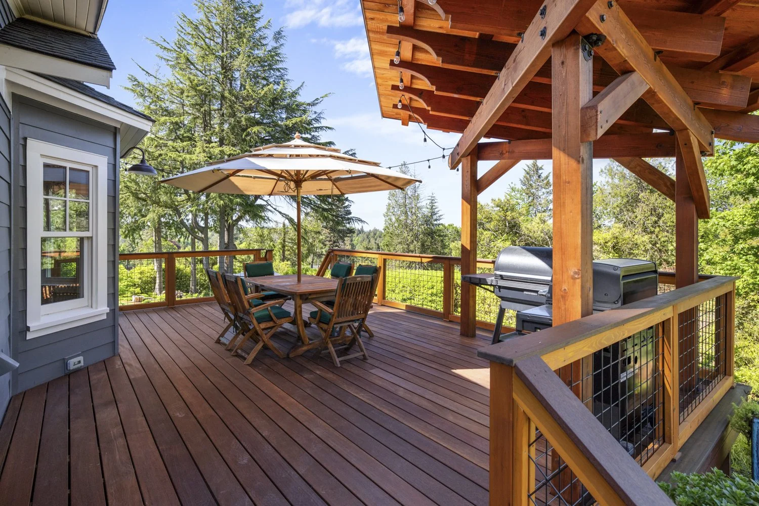 A spacious wooden deck with outdoor dining furniture including a table, six chairs, and a large umbrella. There is also a charcoal grill under a wooden shelter, and the deck is surrounded by trees under a partly cloudy blue sky.