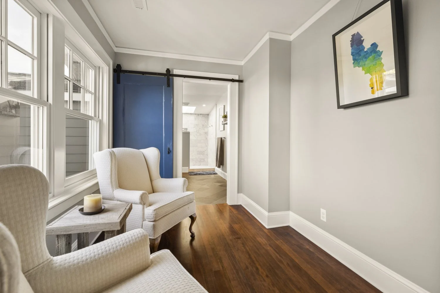 A corner of a living room with two cream-colored armchairs, a small side table with a candle, large windows letting in natural light, and a framed colorful ice cream cone art on the wall. In the background, a doorway with a blue sliding barn door lea