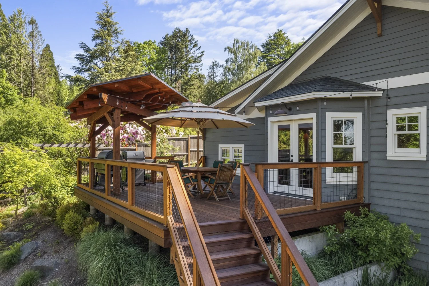 Wooden outdoor deck with railing, stairs, a patio table with chairs, umbrella, grill, and shaded structure, attached to a gray house with white-framed windows, surrounded by greenery and tall trees.