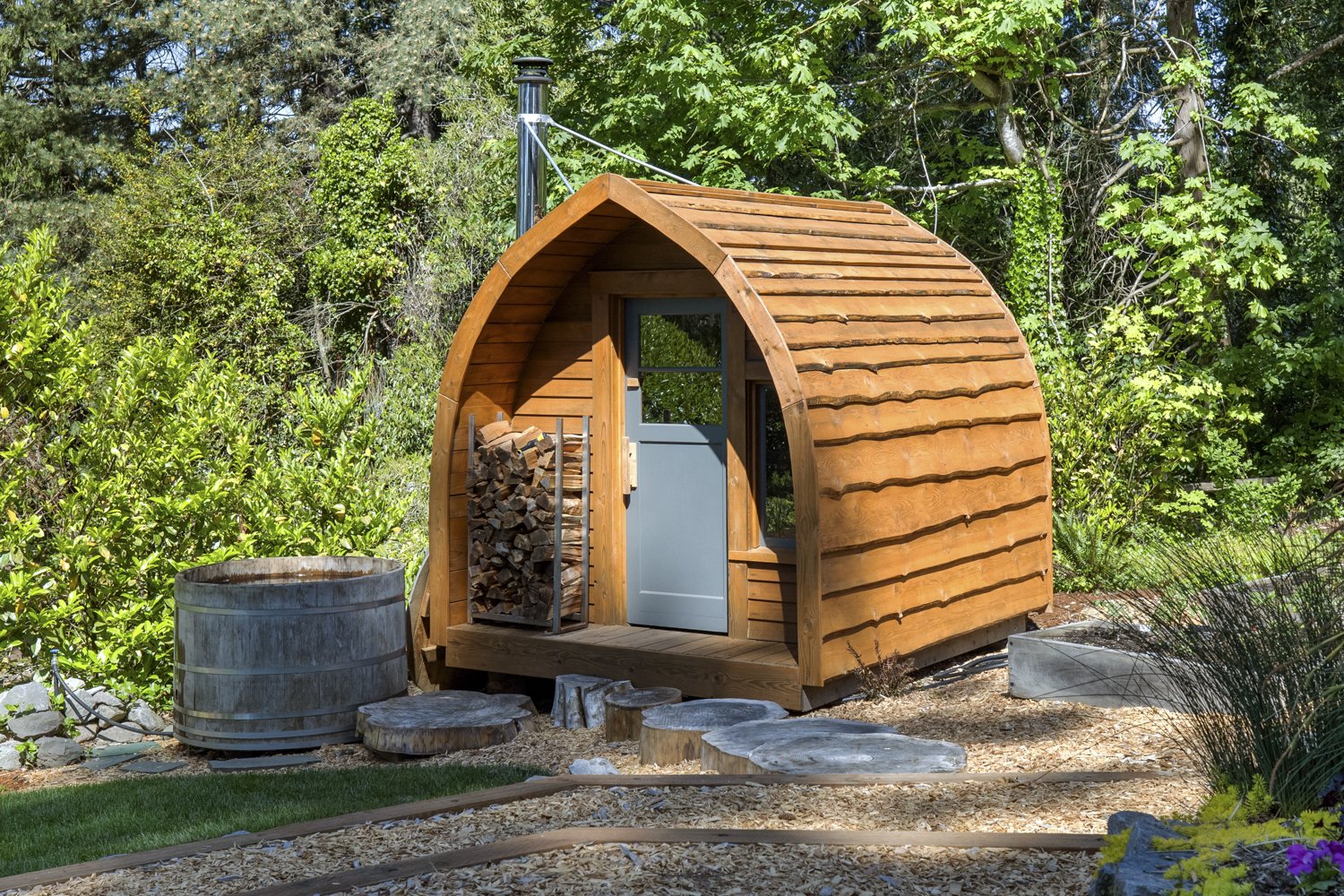 A small, rounded wooden cabin in a garden, with a blue door, wood pile, and surrounding greenery.