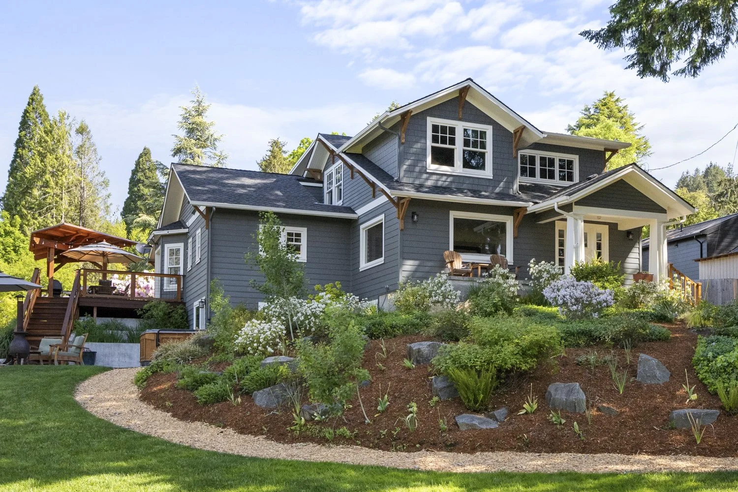 A two-story house with gray siding, white trim, and multiple windows, surrounded by a landscaped garden with various plants, rocks, and a curved pathway. There is a porch with outdoor furniture and a wooden deck with a patio umbrella, set against a b