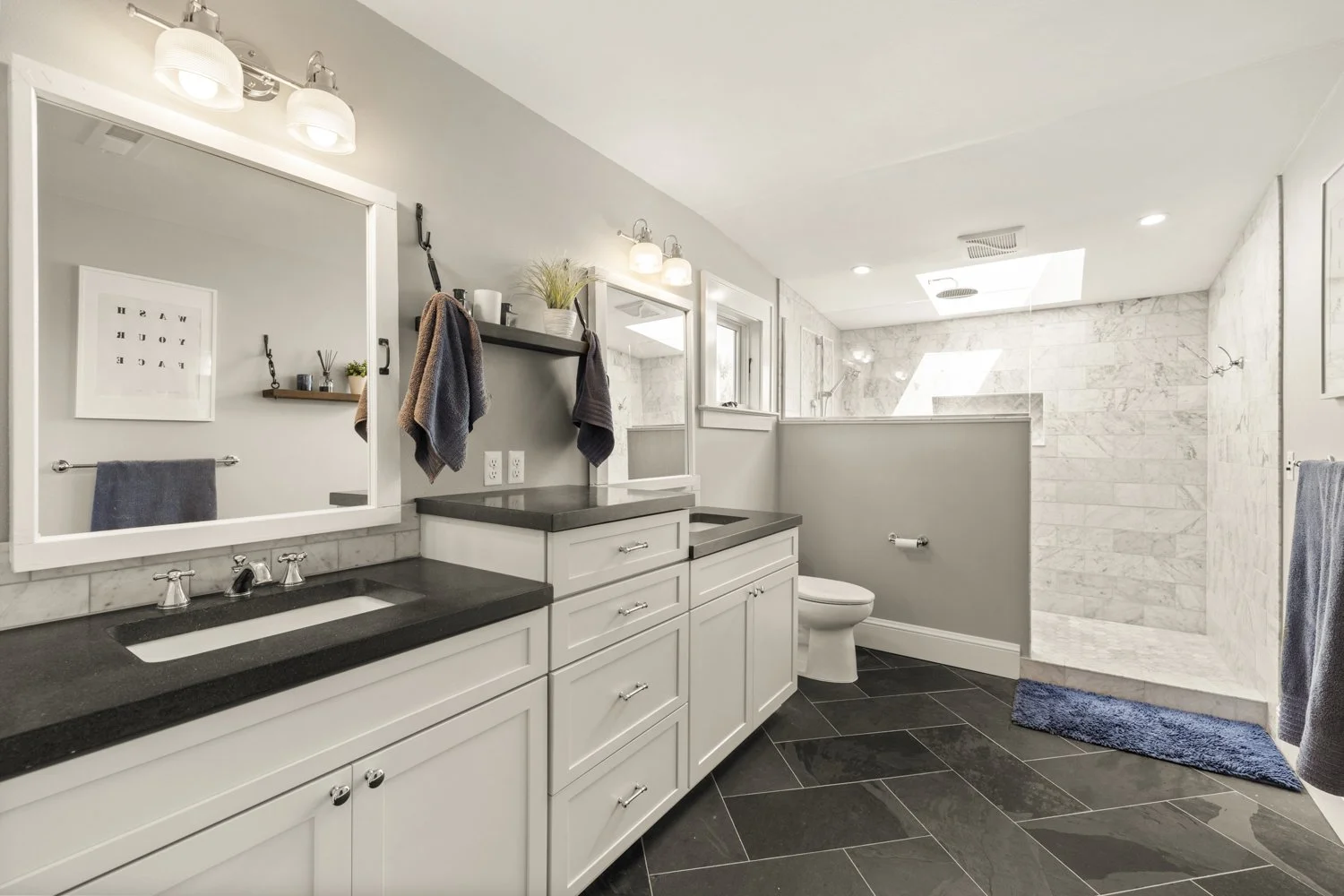 Modern bathroom with double vanity, black countertops, white cabinets, large mirror, towels, and a walk-in shower with marble walls and skylights.