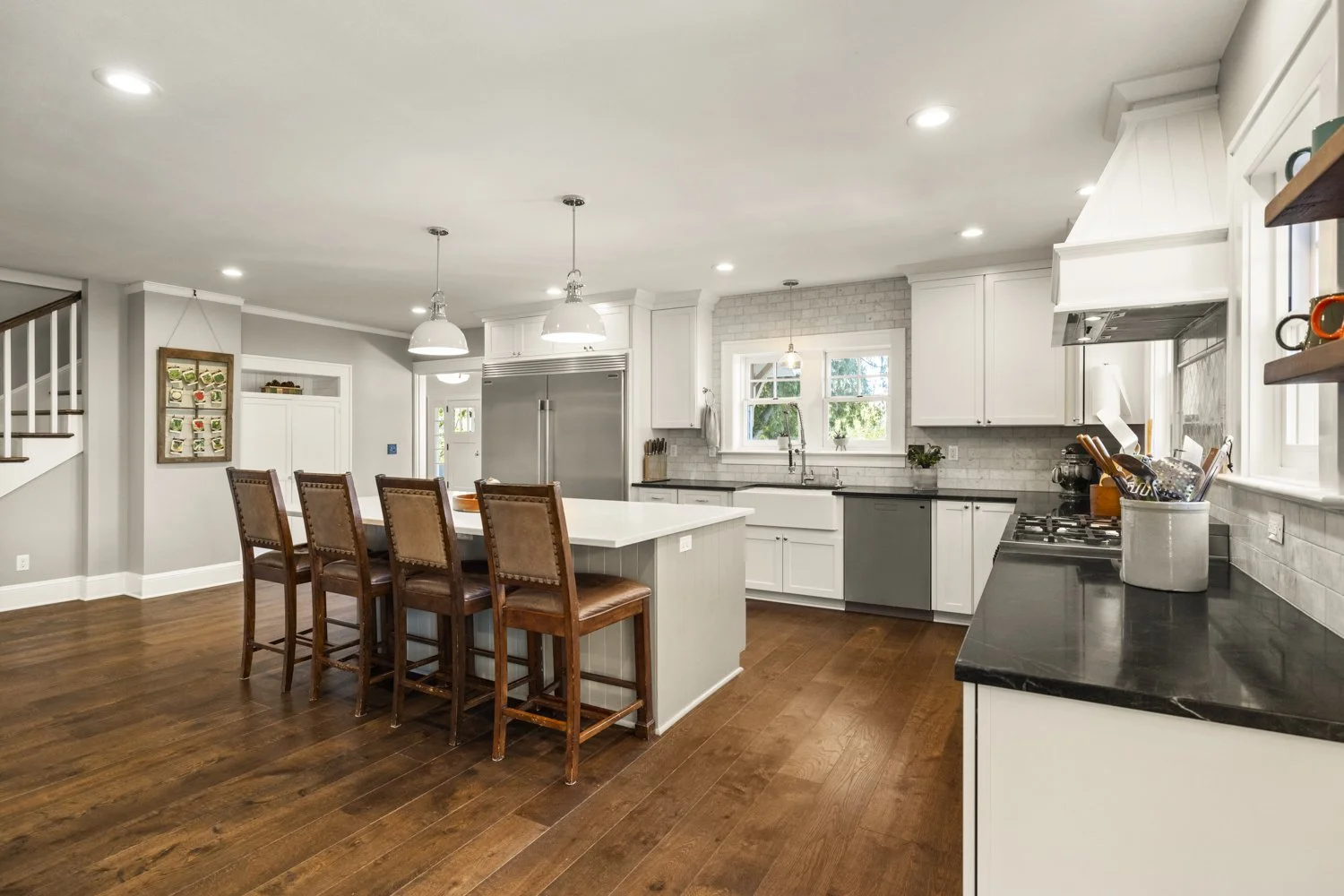 Modern kitchen with white cabinets, black countertops, and wooden flooring, featuring a central island with four brown leather chairs, stainless steel appliances, and a window above the sink.