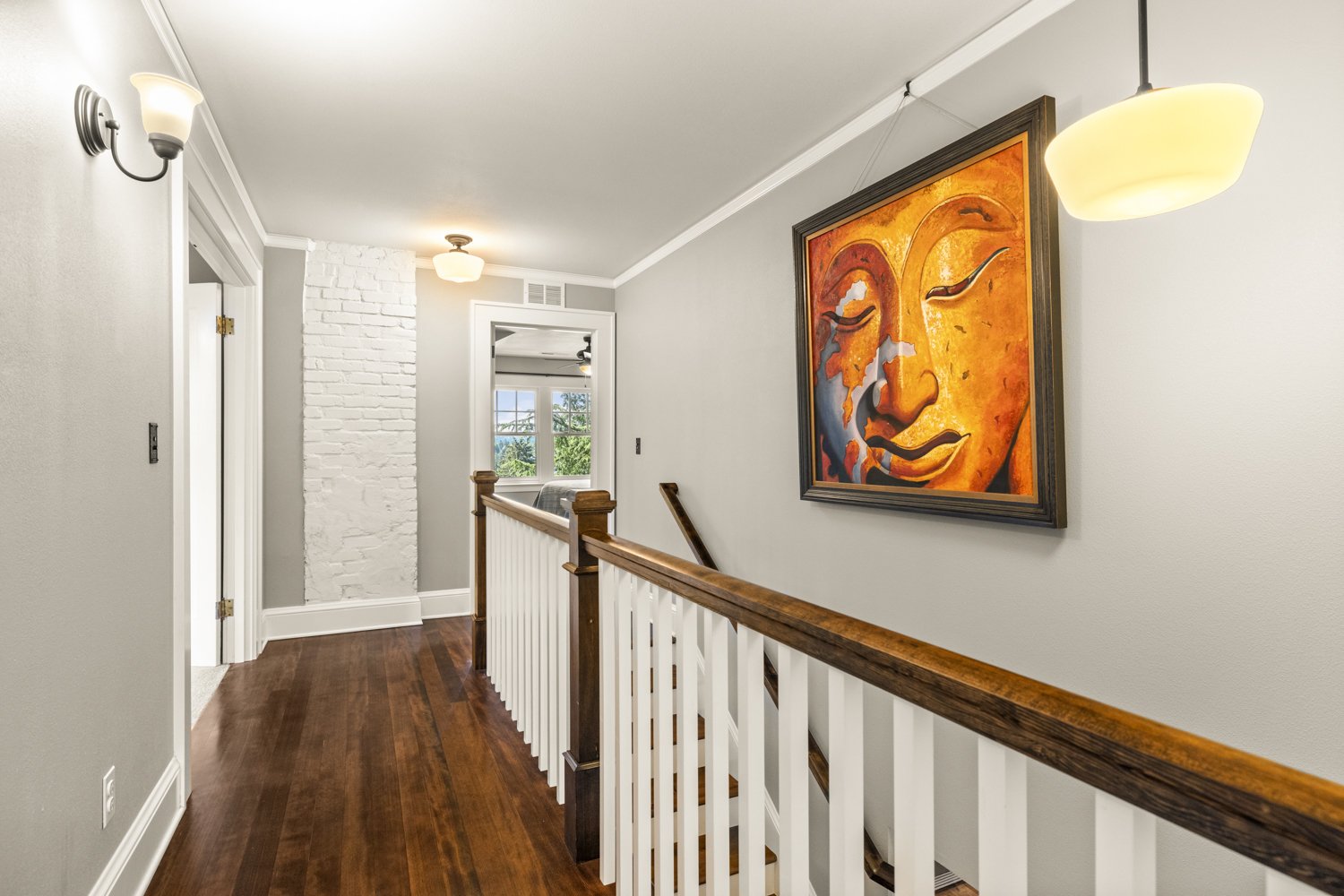Interior of a hallway with hardwood floors, grey walls, a white ceiling, a white brick wall section, framed art of a face, and light fixtures.