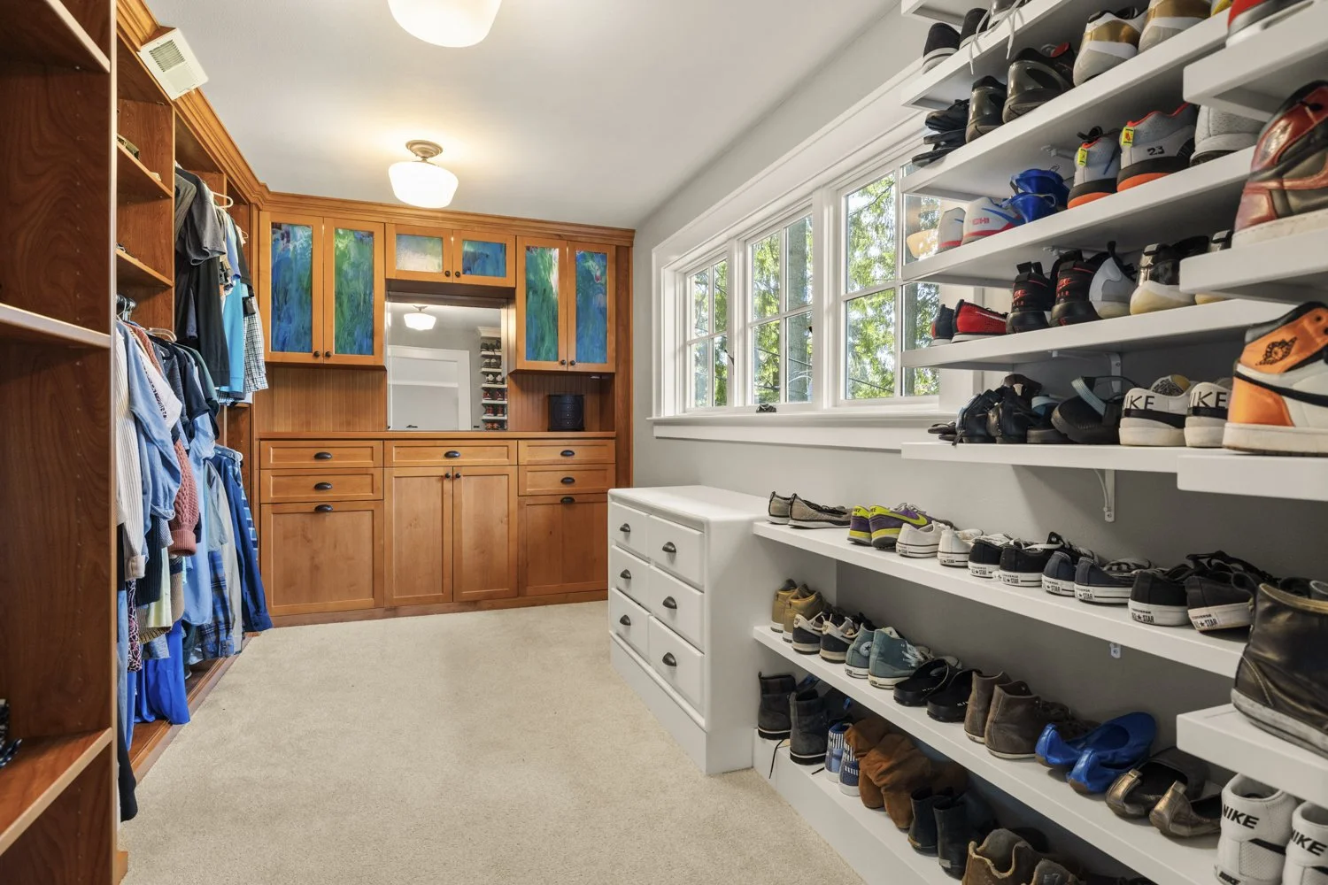 A walk-in closet with wooden cabinets, shelves, and drawers, containing clothes and shoes. There is a large window letting in natural light and a mirror on the back wall.