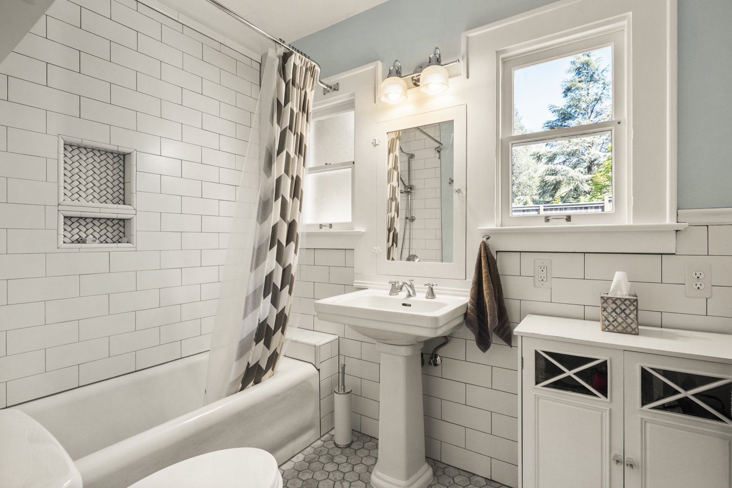 Bathroom with white subway tile walls, a bathtub with a patterned shower curtain, a pedestal sink with a mirror and light fixture, a small cabinet, and windows showing outdoor trees.