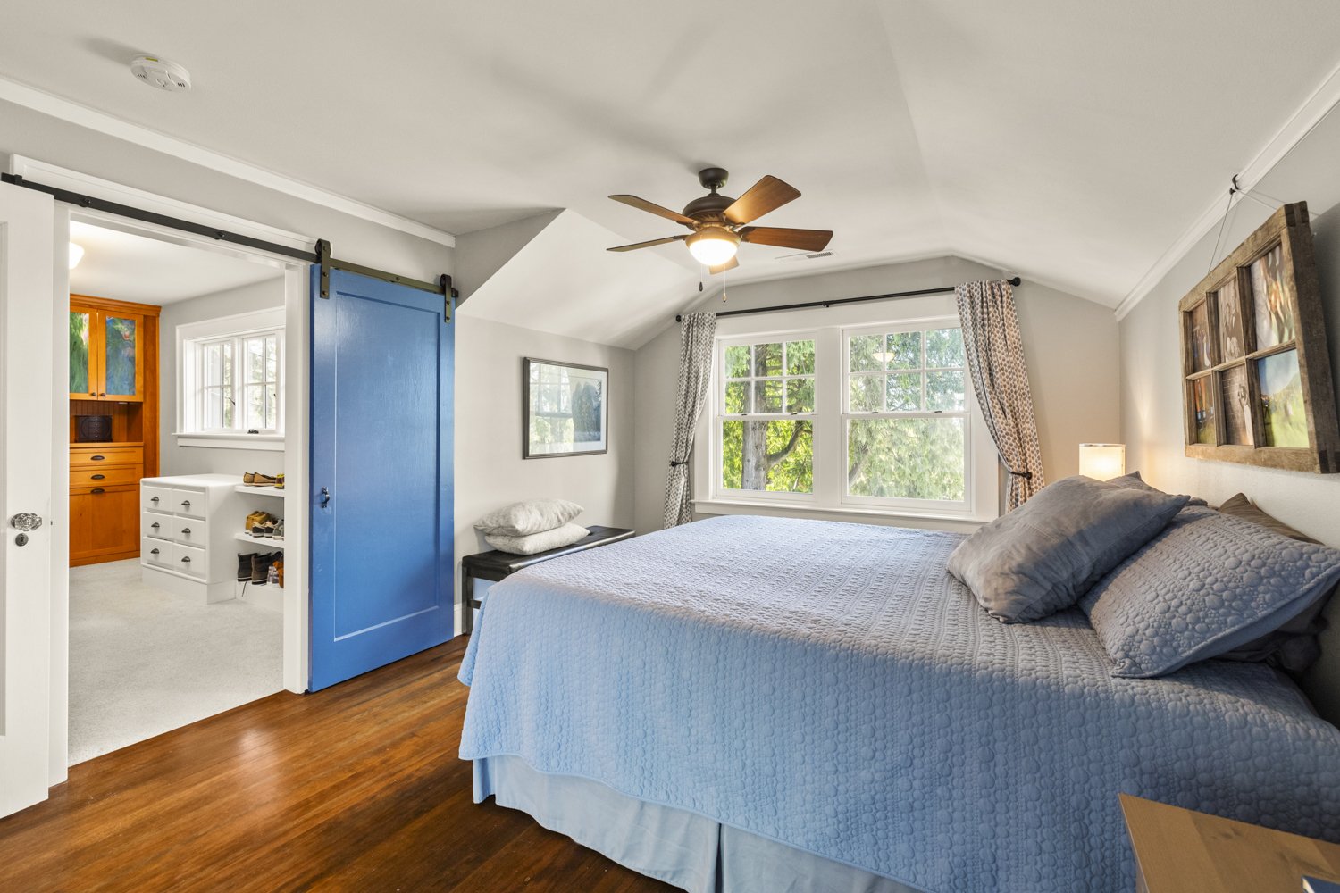 Bright bedroom with window view of trees, large bed with blue quilt, pillows, ceiling fan, and sliding barn door leading to walk-in closet.