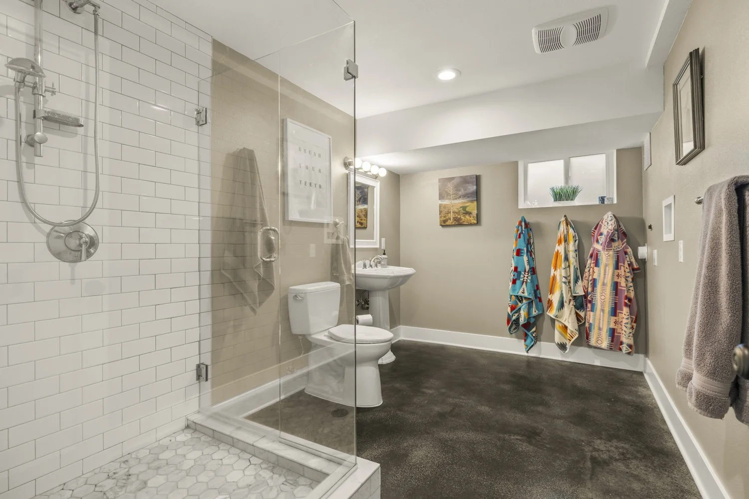 A modern bathroom with a glass-enclosed shower, a white toilet, a pedestal sink with a mirror and light fixture, beige walls, and a dark carpeted floor. There are colorful towels hanging on hooks, a small window with a plant, and framed artwork on th
