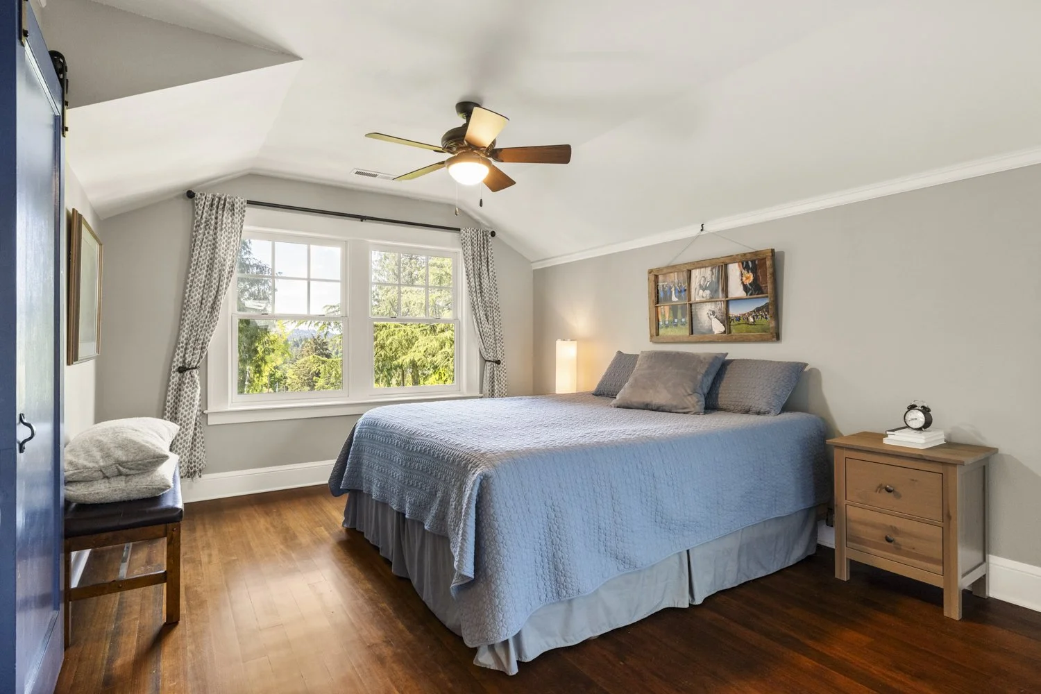 Bedroom with a large bed, wooden side table, window with curtains, wooden floor, ceiling fan, and wall art.