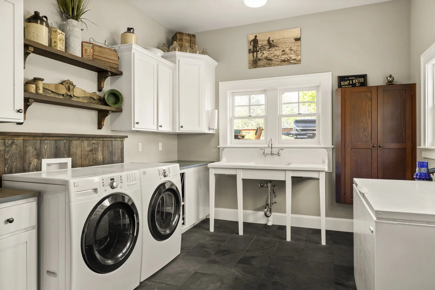 Laundry room with a front-loading washer and dryer, white cabinets, a wooden open shelf with decorative jars, a laundry sink with a window above, and wood and white furniture.