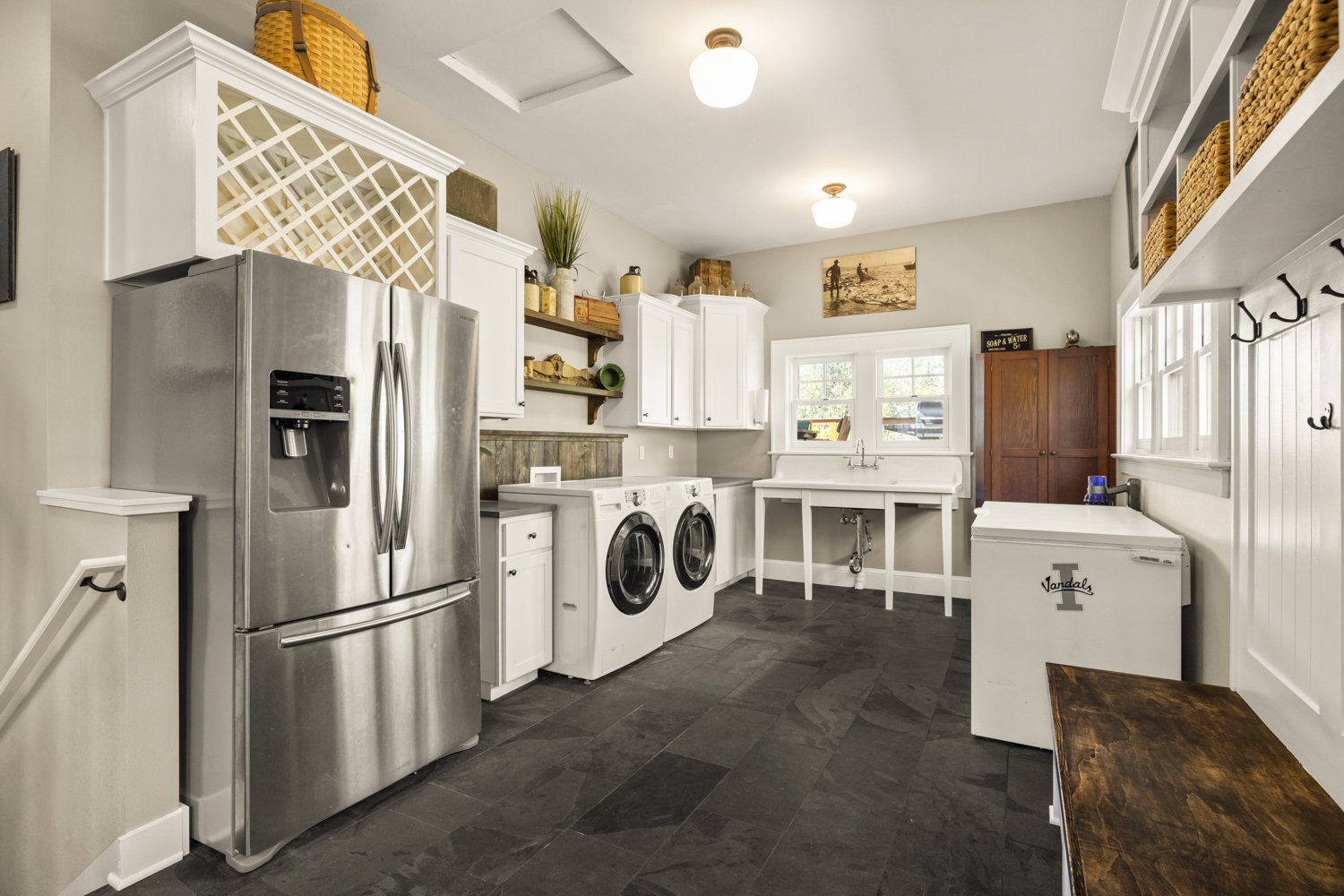Laundry room with stainless steel refrigerator, white washer and dryer, utility sink, open shelving with baskets and decor, wooden cabinet, and wall hooks, in a well-lit space with windows.