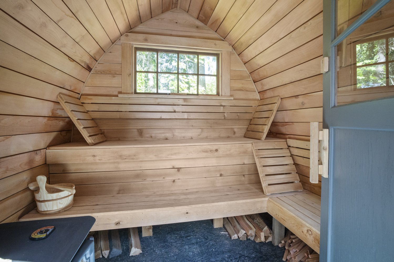 Wood-paneled interior of a small sauna with seating and a small window showing trees outside.