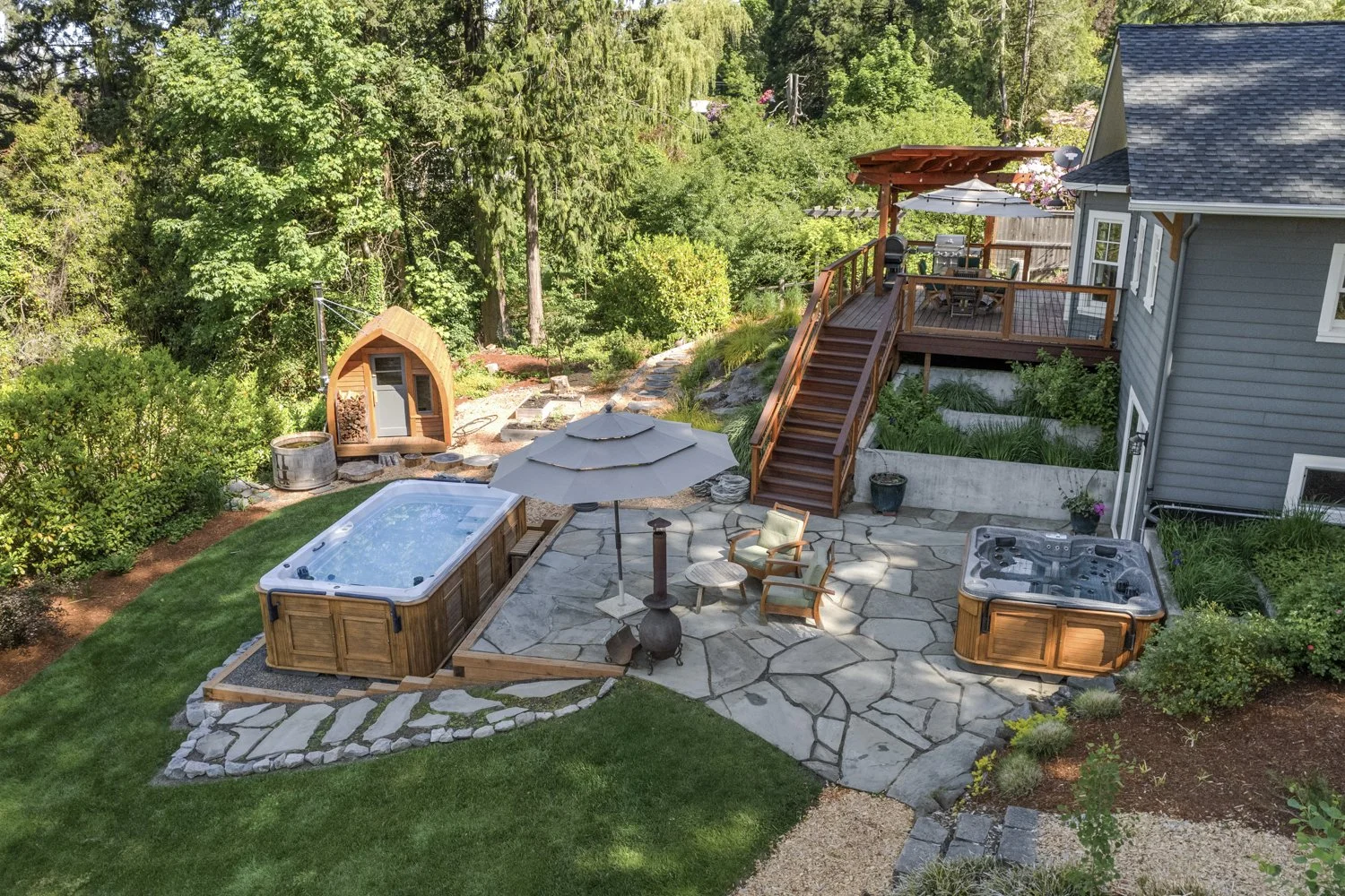 Backyard patio with hot tub, gazebo, seating area with umbrella, hot tub, stairs leading to an elevated deck with outdoor dining, hot tub, and grill, surrounded by lush greenery and trees.