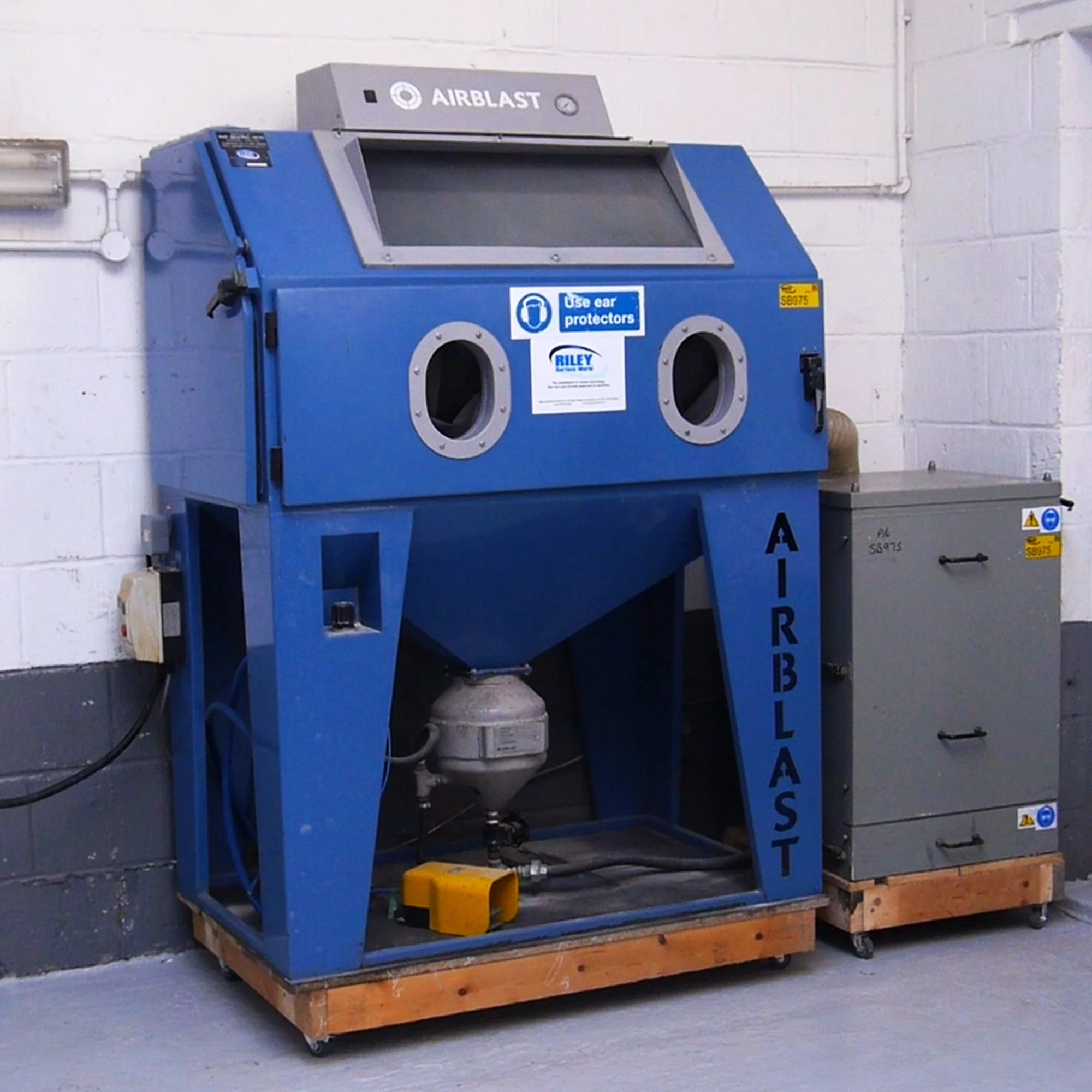 Blue air blasting machine with two circular openings, labeled 'AIRBLAST,' against a white brick wall, with electrical components and a gray cabinet nearby.