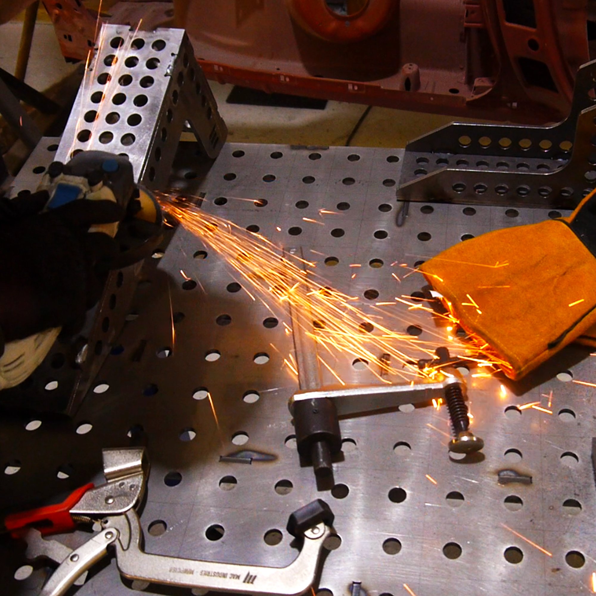 A welding work in progress on metal parts in a workshop, with sparks flying as a person uses a welding torch, wearing gloves and a protective glove on one hand.