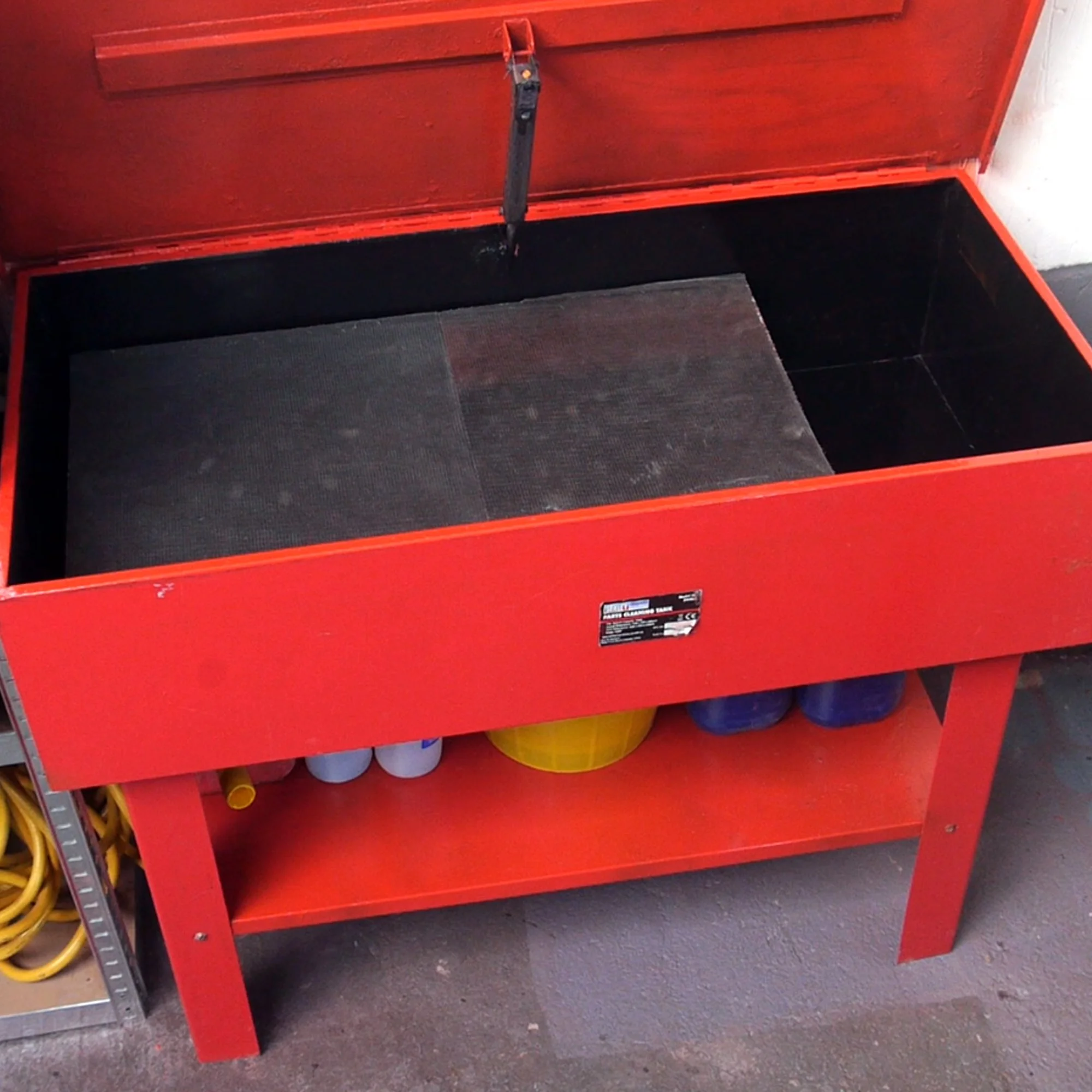 Red workbench with closed top, underneath has a lower shelf with yellow and blue plastic containers, and an empty black top tray.