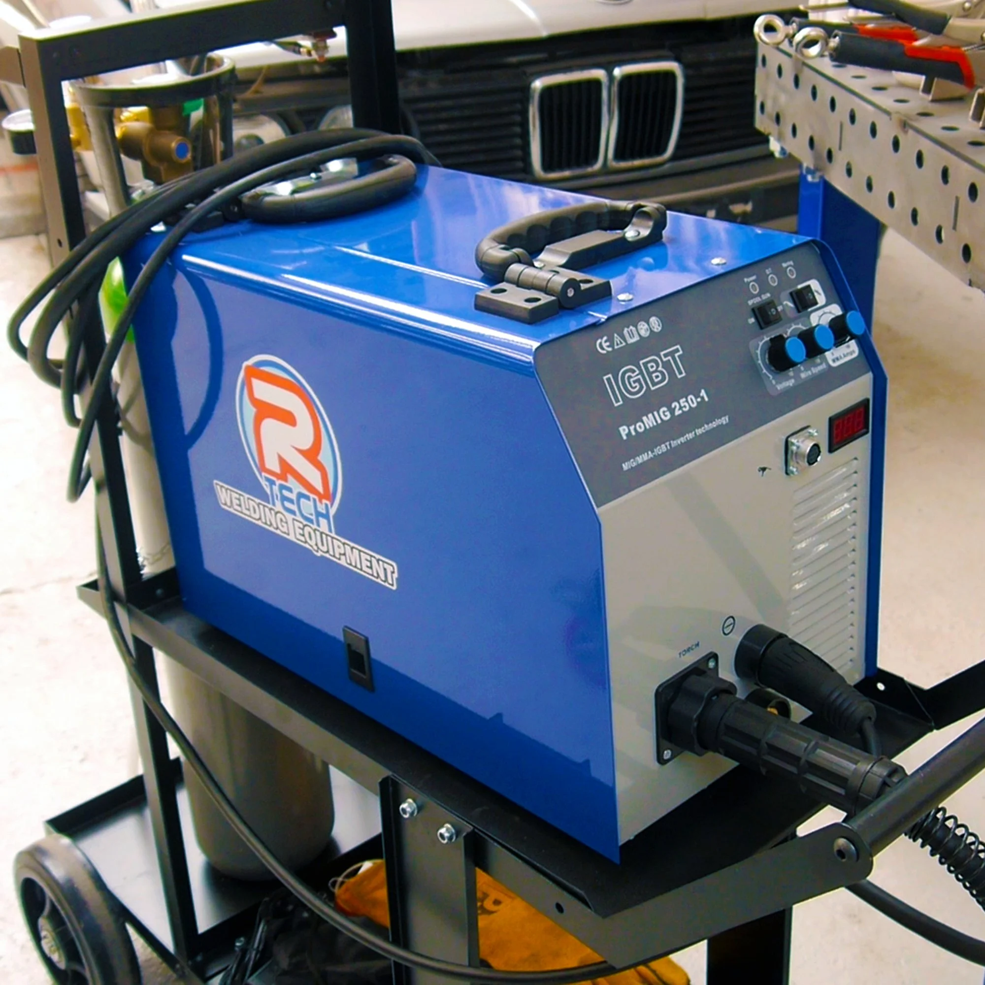 Blue welding machine with controls and hoses, labeled R TECH welding equipment, situated on a wheeled cart in a workshop.