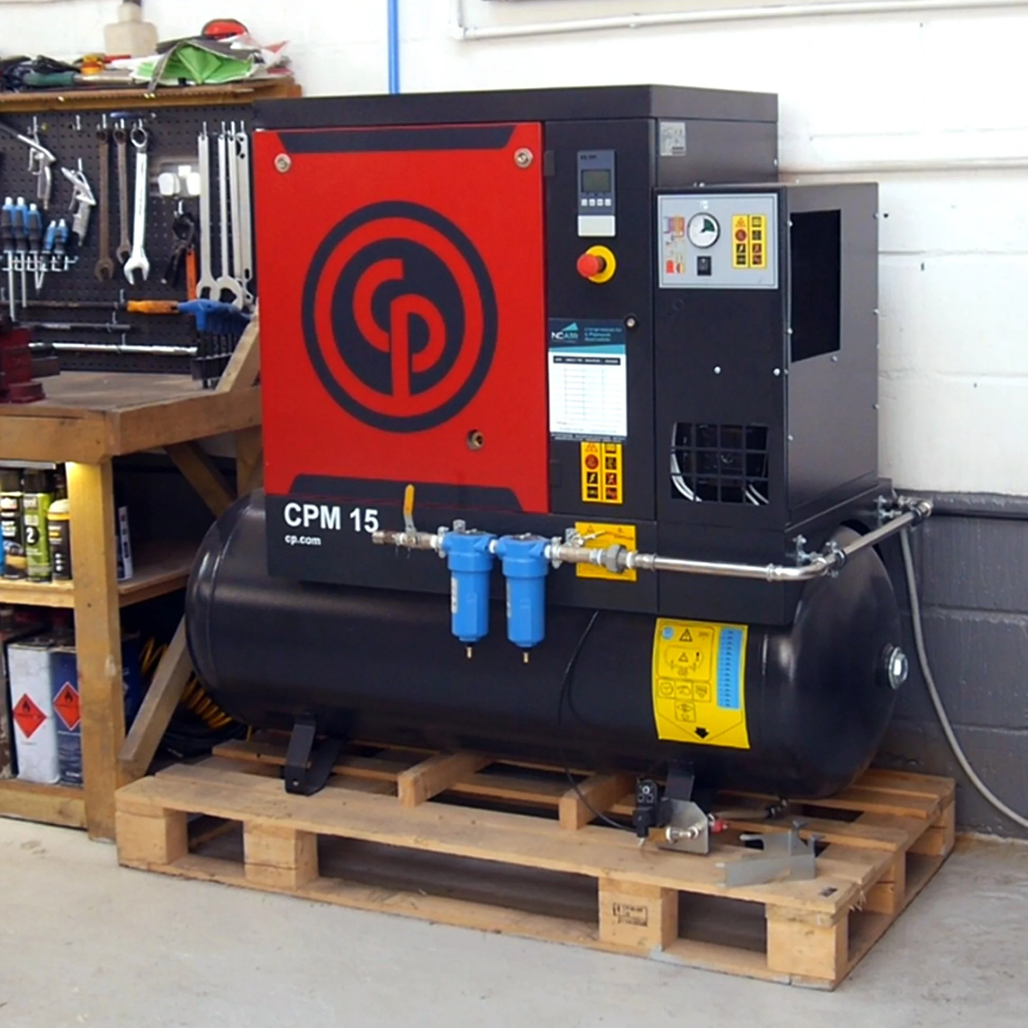 Industrial air compressor with black tank, red and black control unit, and blue water filter, placed on wooden pallet in workshop.