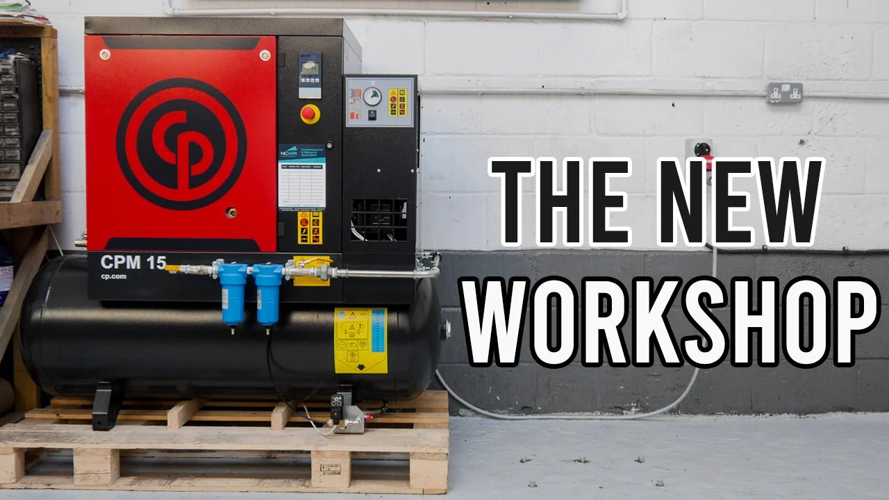An industrial workshop with a large air compressor machine on a wooden pallet, positioned against a gray and white brick wall, with the text 'The New Workshop' overlaid on the right side of the image.