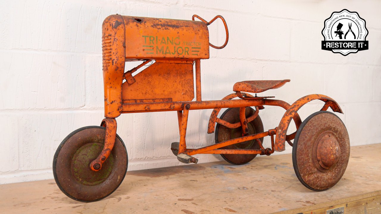 A vintage orange tricycle with a rectangular seat and a large box labeled 'TRIANG MAJOR'. The tricycle appears rusted and weathered, displayed against a white brick wall background.