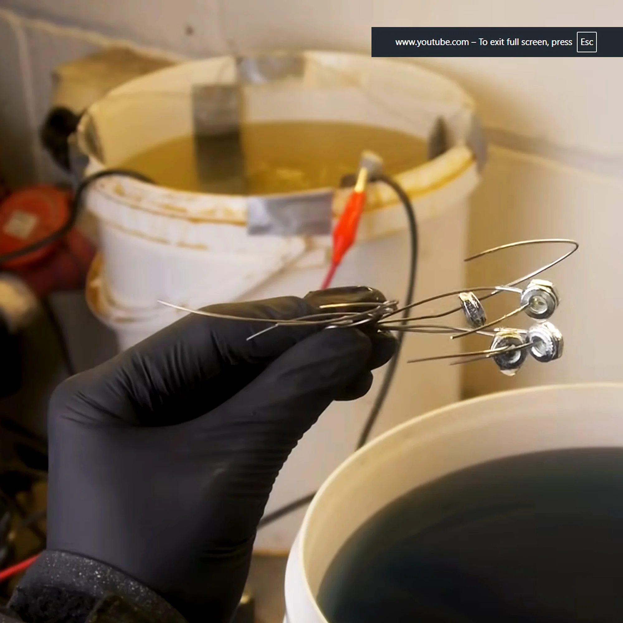 A person wearing a black glove holds a small circuit with three large metal components and wires attached, with a large container of dark liquid in the background.