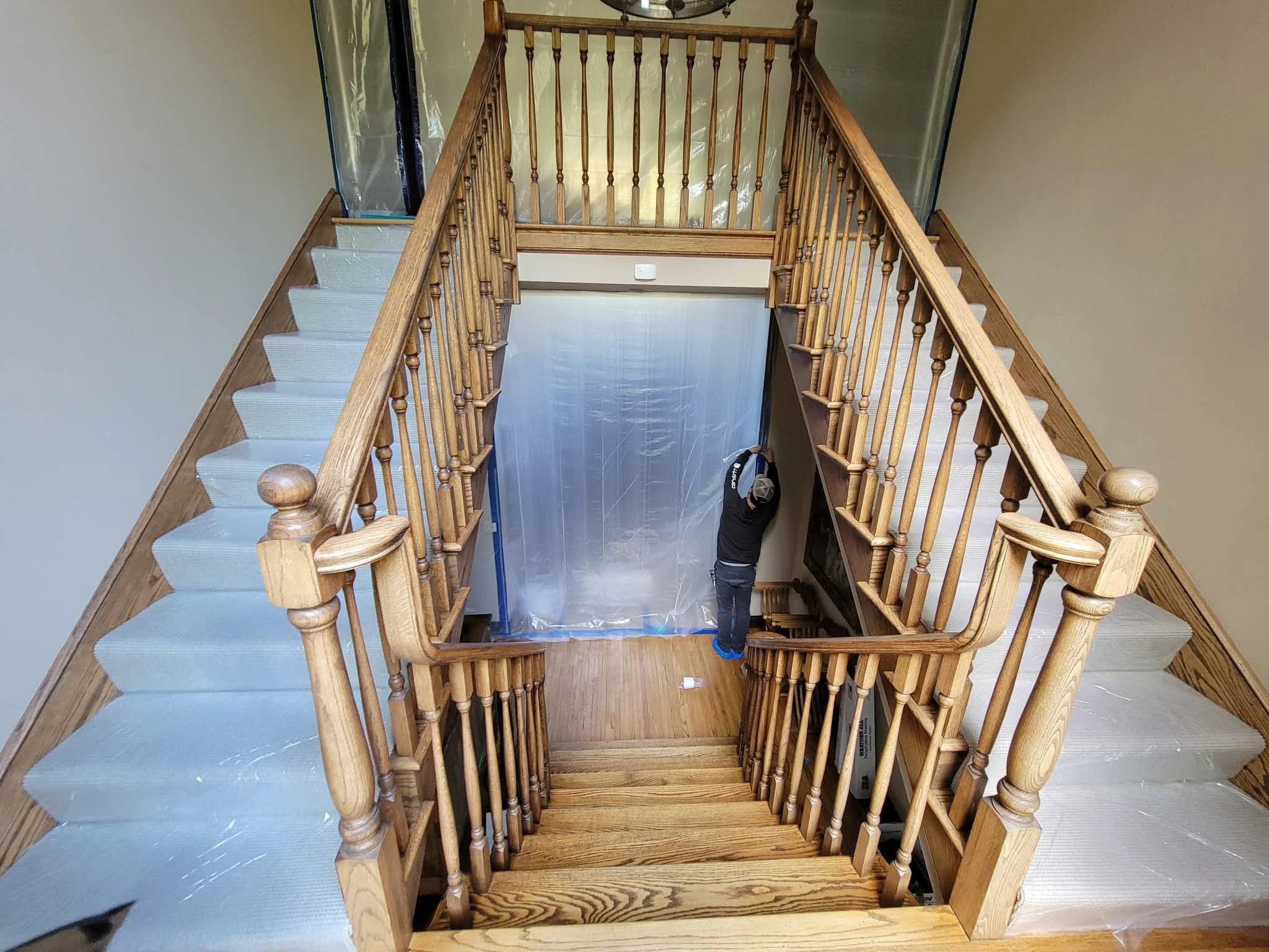 A staircase with wooden railings and beige carpeting, leading to upper and lower floors. A person is standing at the bottom, facing a plastic sheet covering a doorway or opening.