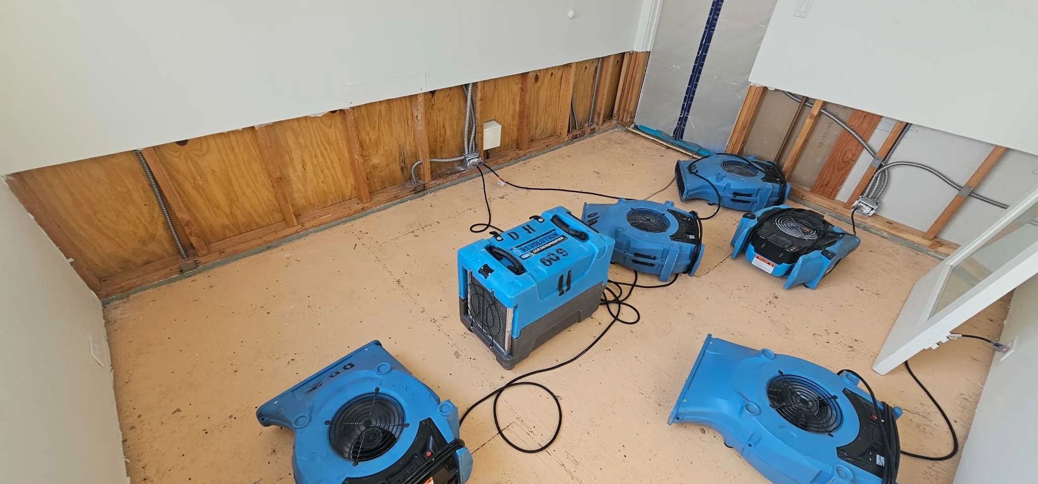 Room under renovation with exposed wooden wall and ceiling, electrical wiring, and multiple blue portable air movers on the floor.