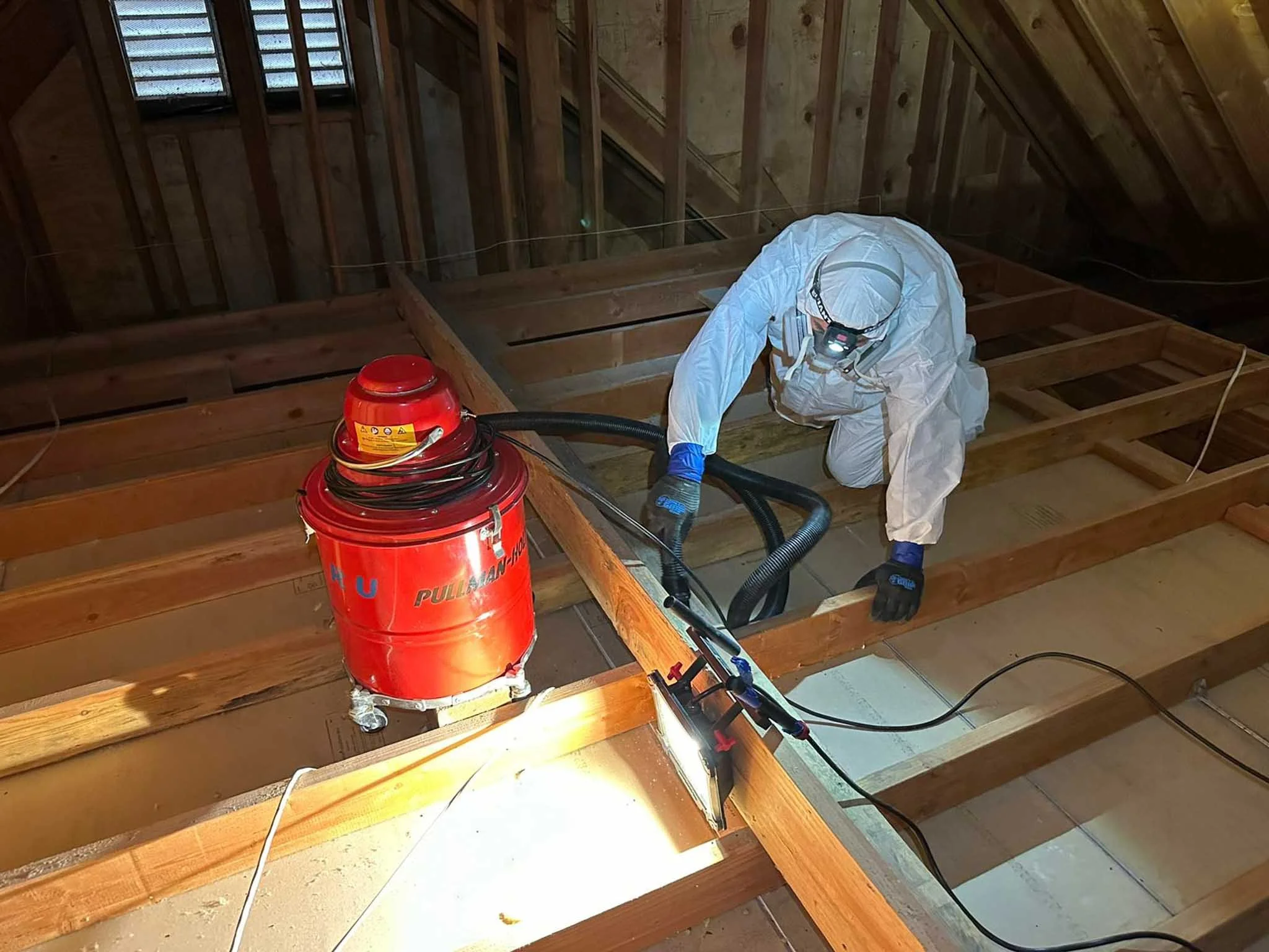 A worker dressed in protective gear, including a white coverall, gloves, and a headlamp, is working on a wooden floor frame in a construction site. The worker is using a vacuum or extraction device, connected to a red industrial vacuum, to possibly remove dust or debris from the wooden structure.
