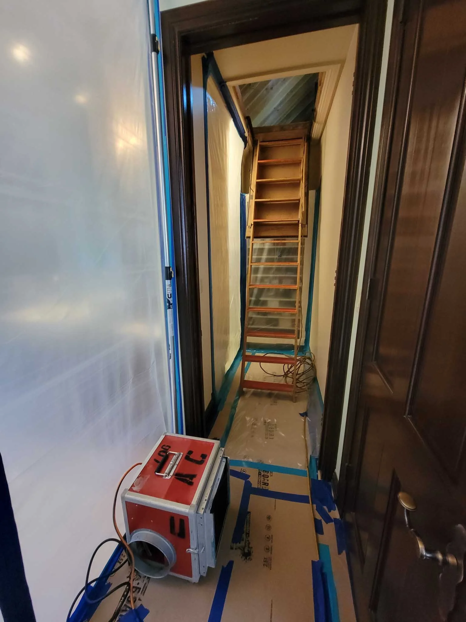 A narrow hallway under construction, with a retractable attic ladder extended into the ceiling, and a portable air scrubber on the floor near the door.