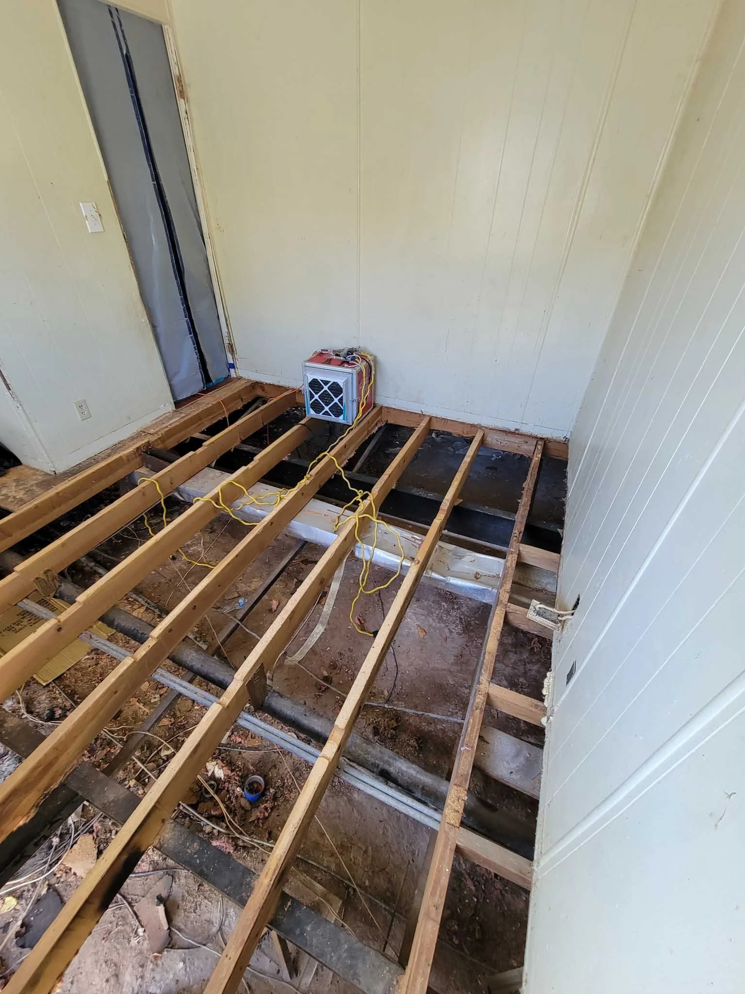 A room with exposed wooden floor joists, some black pipes, and electrical wires. The walls are painted in a light color, and there is a small HVAC unit positioned at the far corner near a doorway.