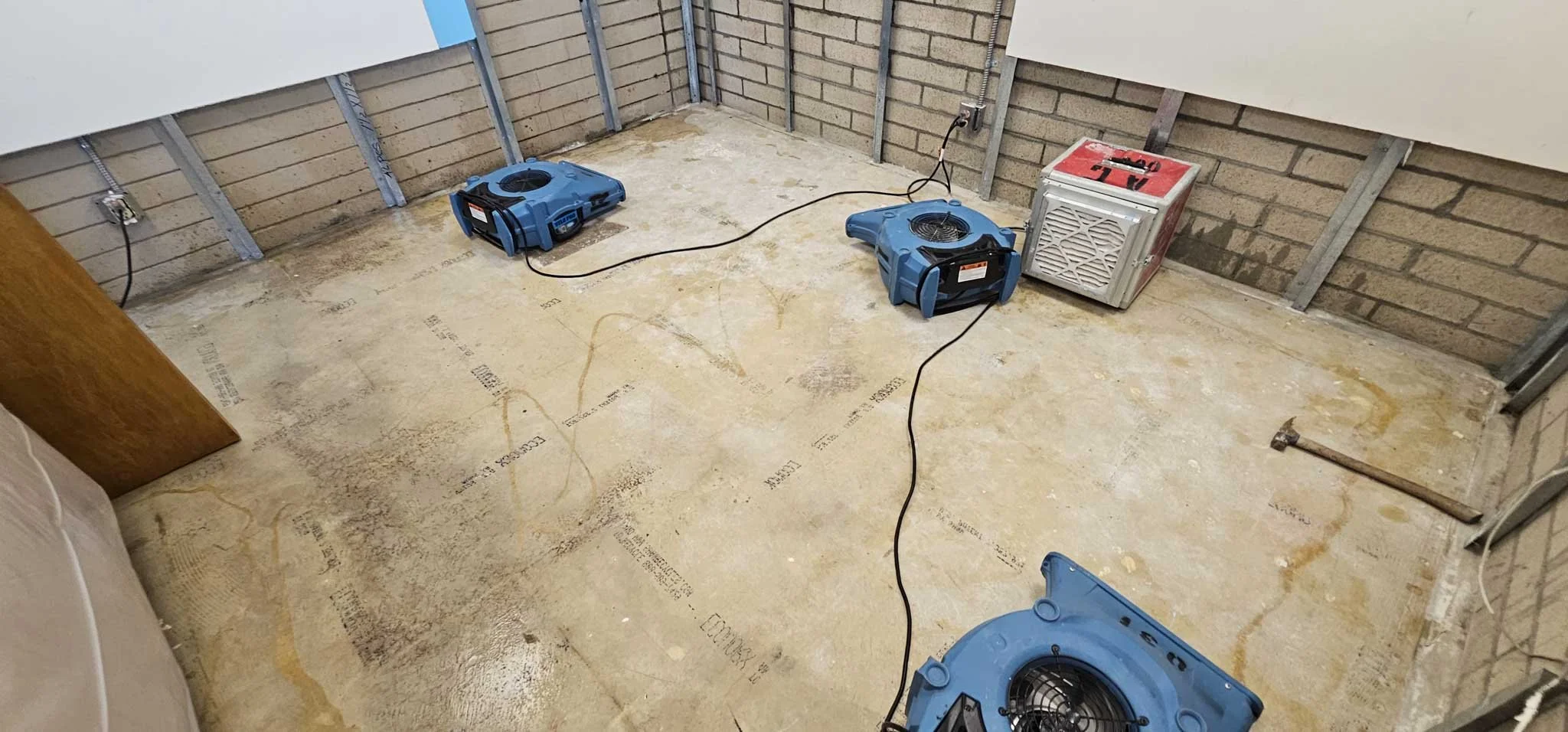 Unfinished room with brick walls and concrete floor, equipped with two portable air movers, a utility axe on the floor, and a mini split AC unit in the corner, indicating ongoing construction or renovation.