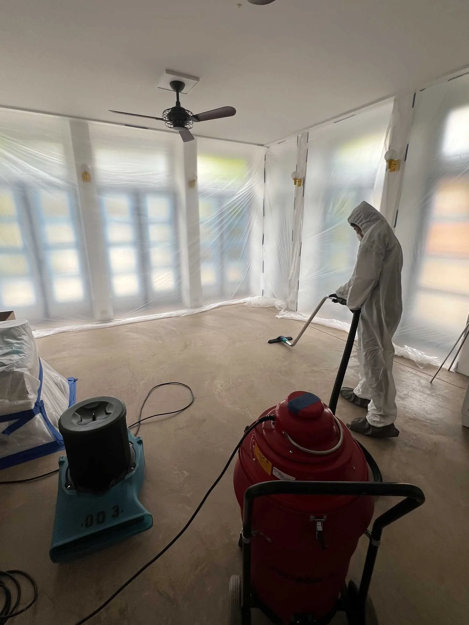 Person dressed in protective suit cleaning or disinfecting an empty room with a vacuum or fogging tool, surrounded by transparent plastic sheeting.