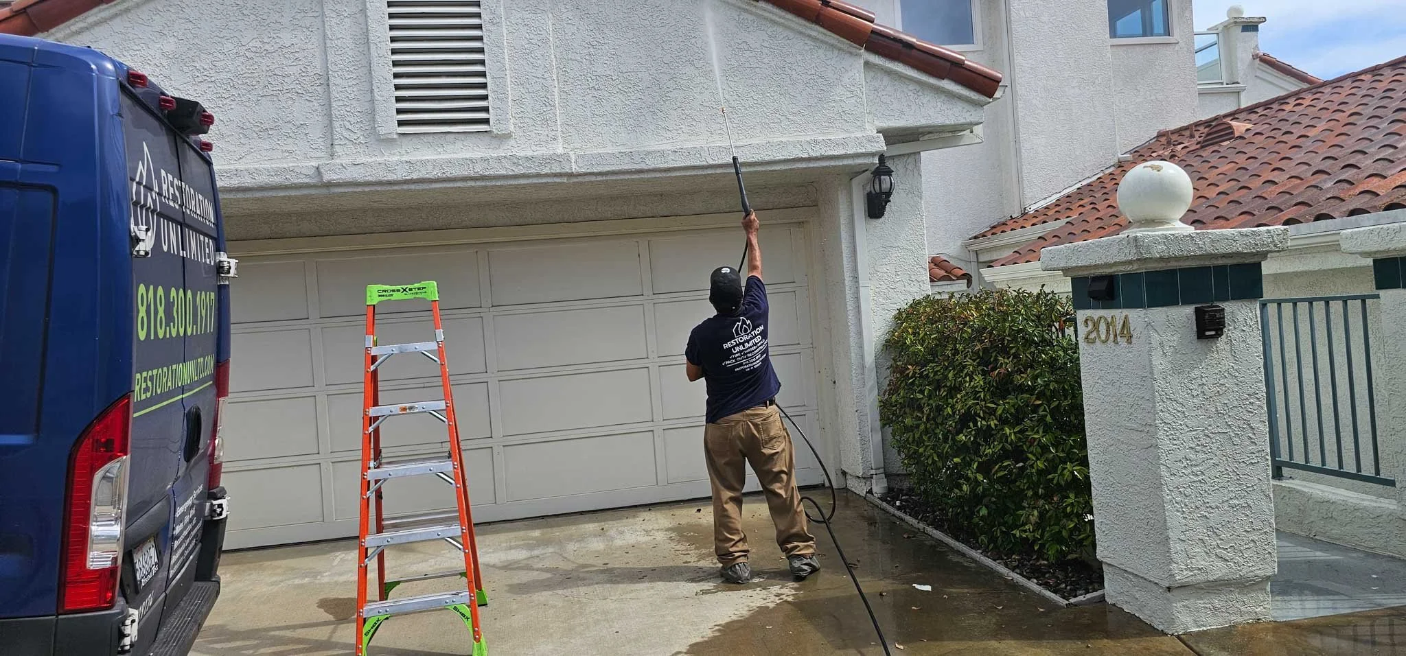 A worker pressure washing a concrete driveway in front of a residential house. The worker is holding a long wand connected to a hose, aiming it at the driveway. There is a orange step ladder and a blue van parked on the left side with company details.