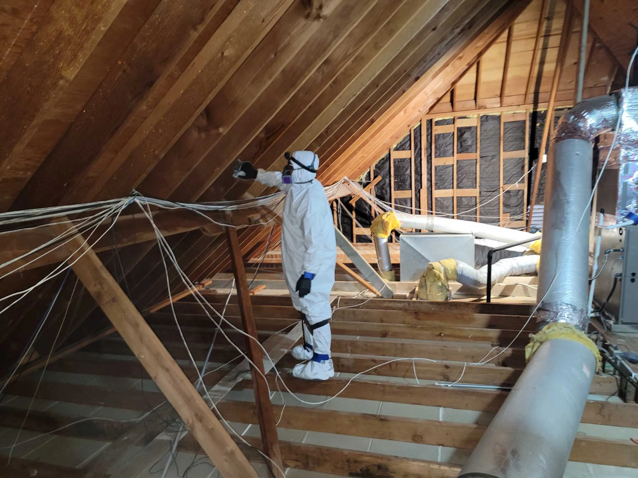 Person in protective suit working on attic insulation and ductwork in a house.