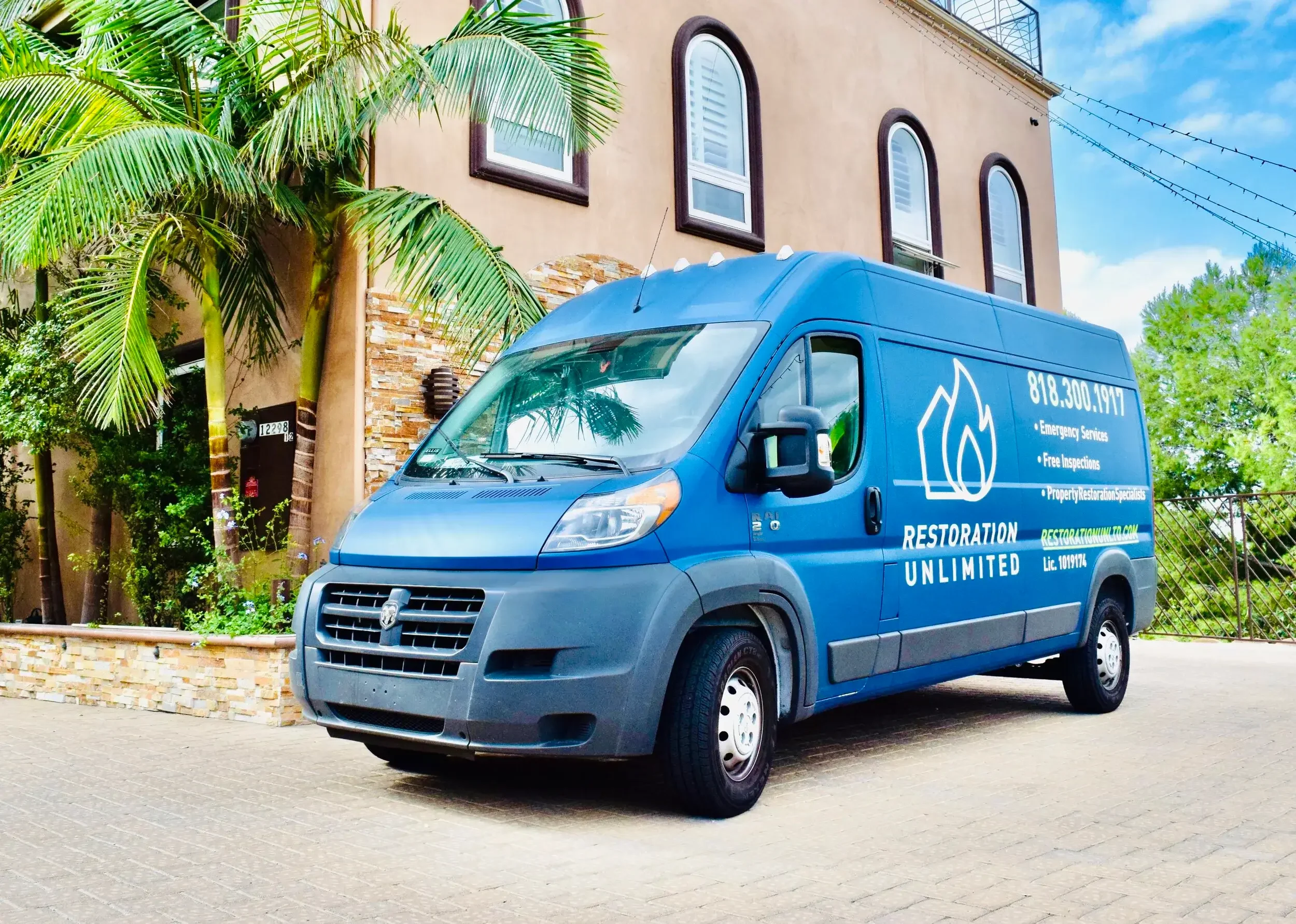 Blue restoration service van parked in front of a beige residential building with palm trees and green foliage.
