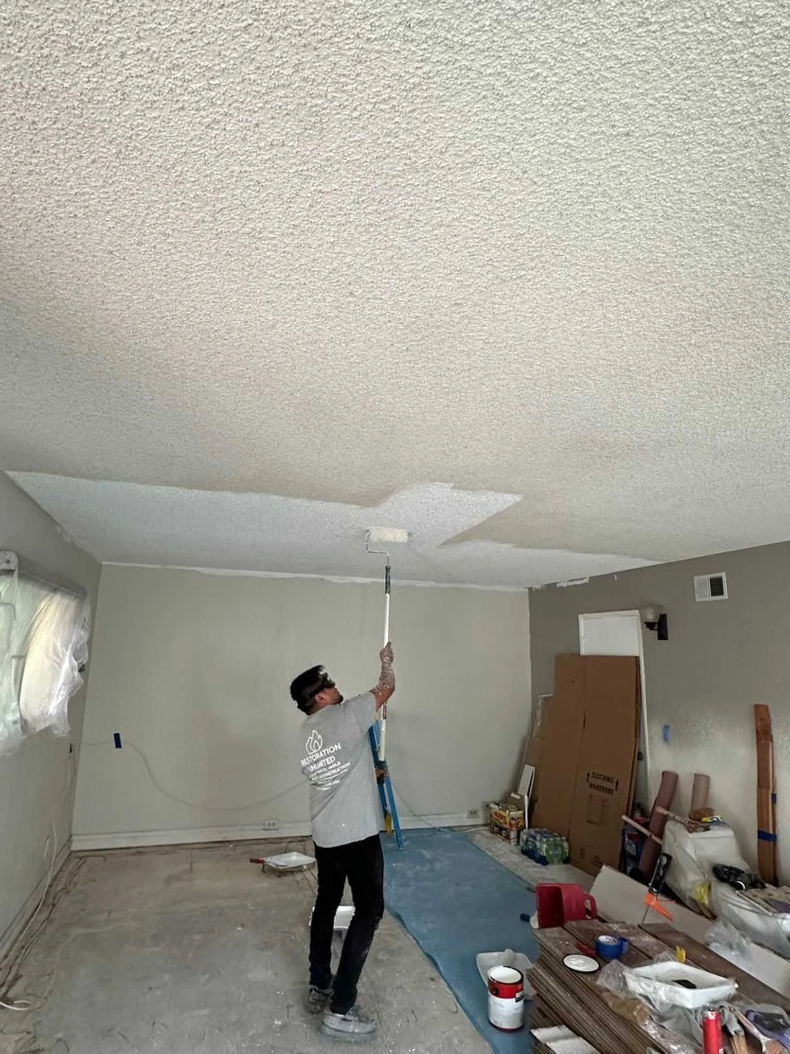 A person painting a ceiling with a roller in a room under renovation. The room has tools, paint supplies, and construction materials scattered around.