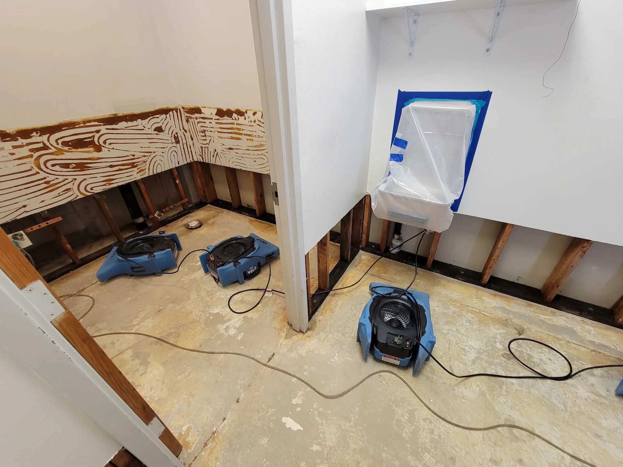 Bathroom construction with a toilet installed, two heater fans on the floor, exposed wall studs, and a plywood wall with wood grain design.