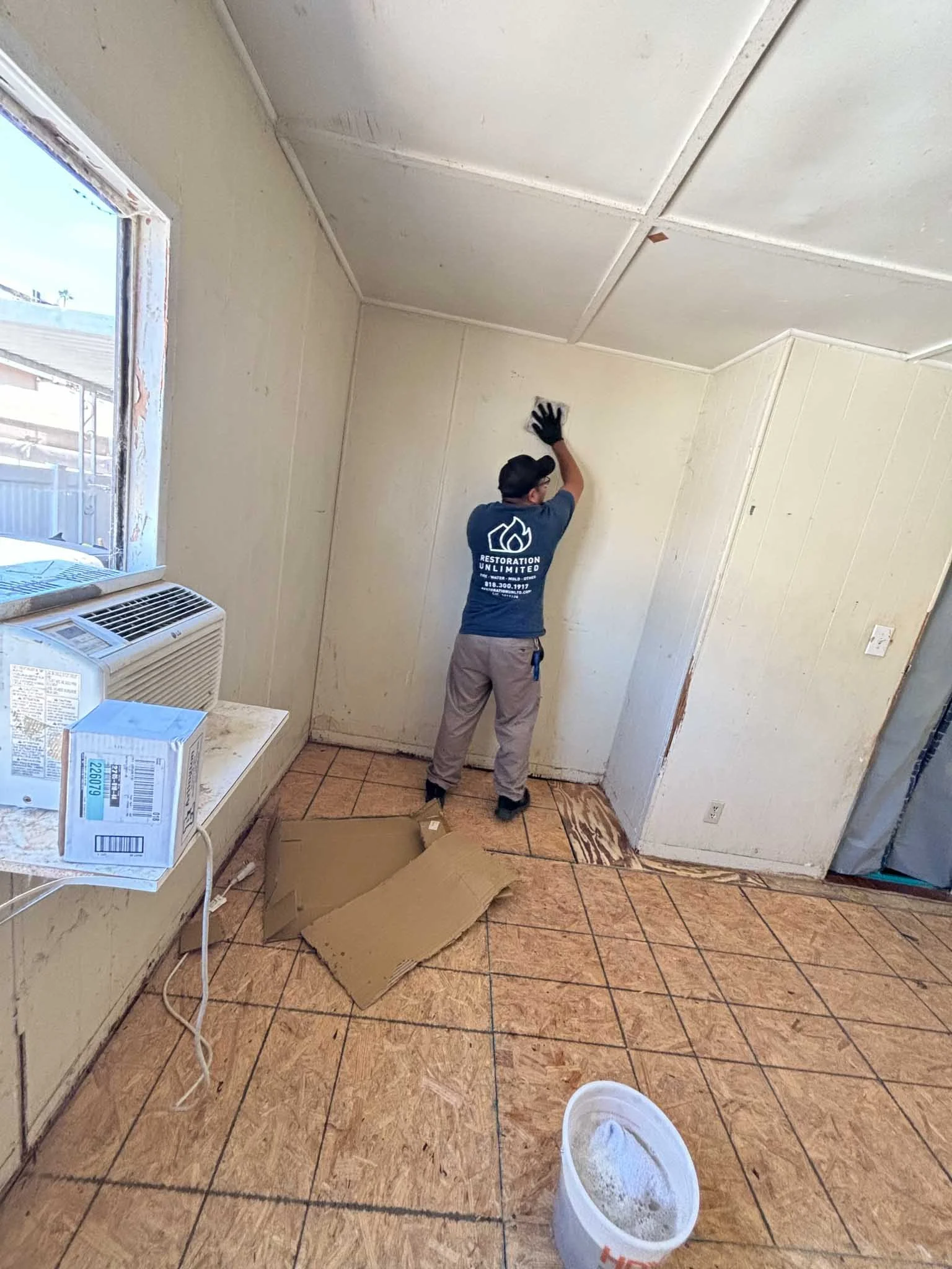 A person in a blue t-shirt and gray pants is cleaning a wall in a small room with beige walls and a partially tiled ceiling. The room has a window with an air conditioning unit and some cardboard pieces on the floor. The person is wearing black gloves and a black cap, and is reaching up to wipe the wall.