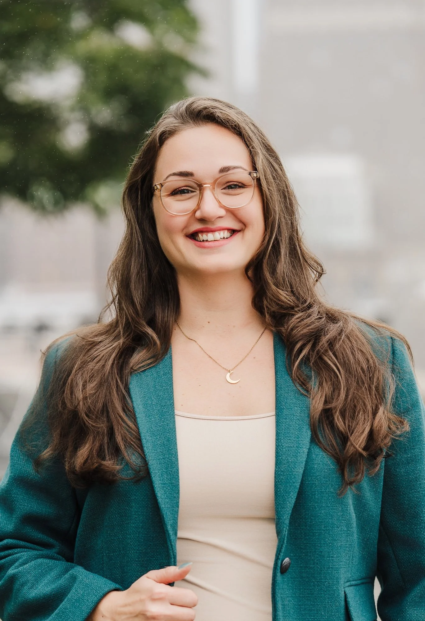 A young woman with long wavy brown hair and glasses, smiling, wears a teal blazer and a gold necklace with a crescent moon pendant, standing outdoors with blurred greenery in the background.
