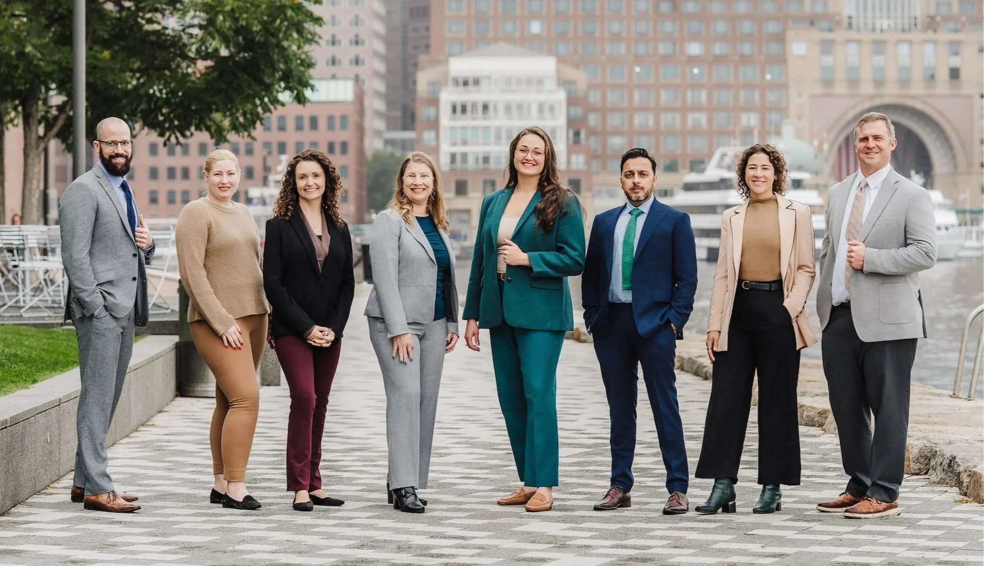 Group of eight diverse businesspeople standing outdoors on a city waterfront, dressed in formal business attire, smiling and posing for the photo.