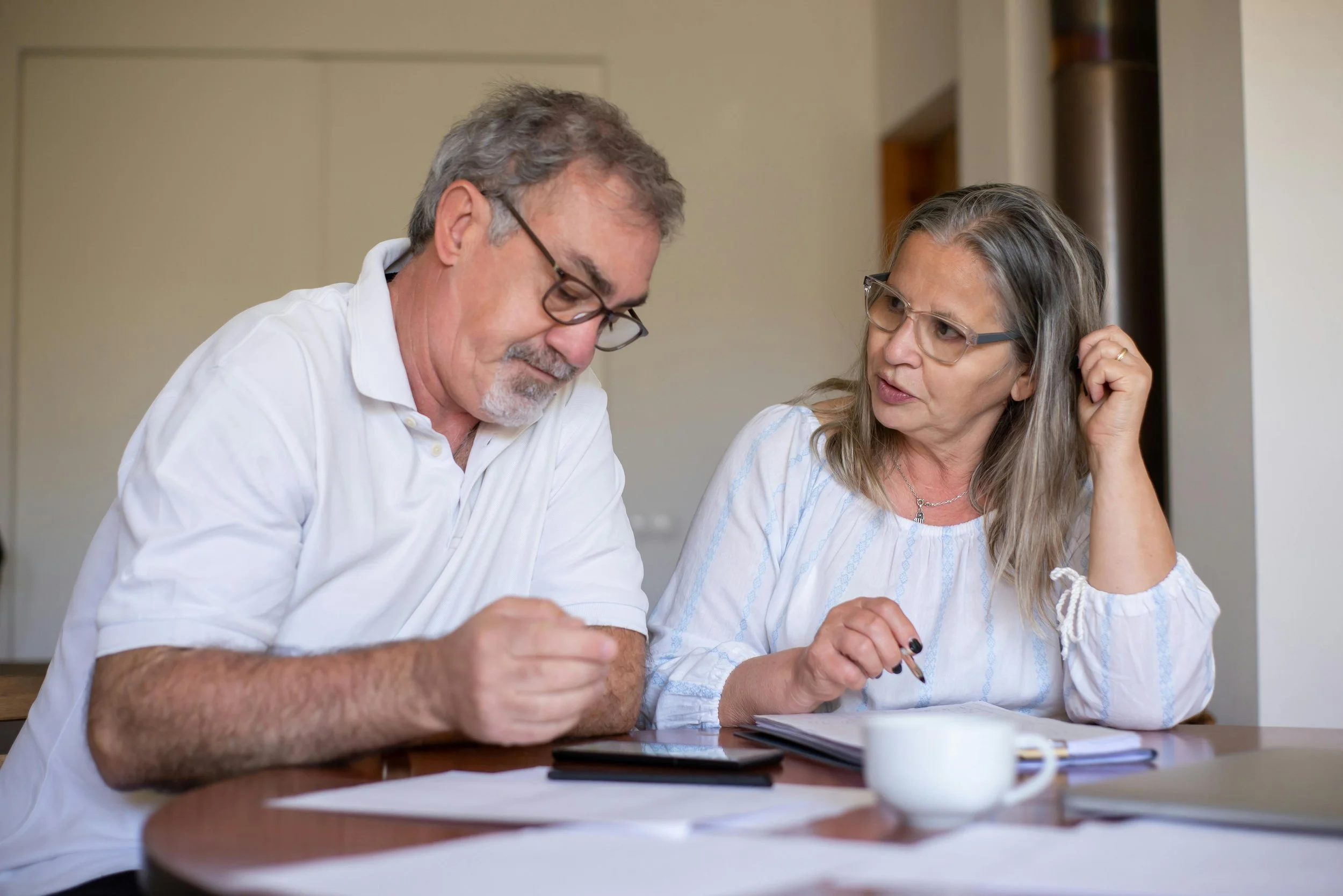 A man and a woman sitting at a table, engaged in a discussion, with papers, a tablet, and a cup in front of them.