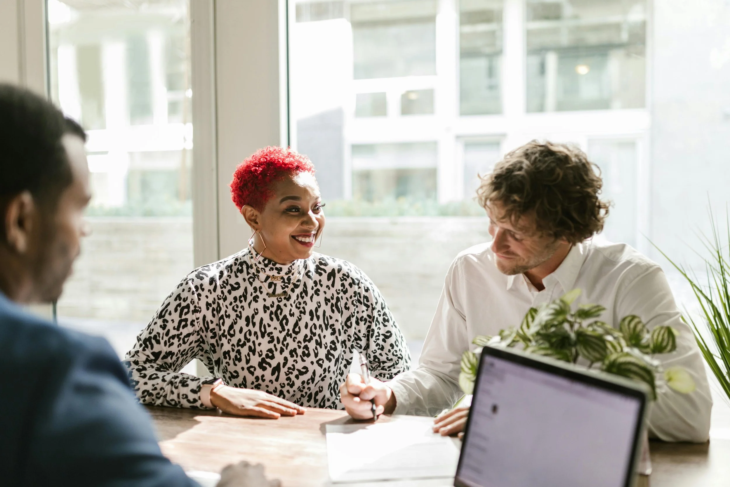 A diverse group of people having a meeting in a bright, modern office with large windows, one woman with red curly hair and leopard print top smiling and engaged in conversation.