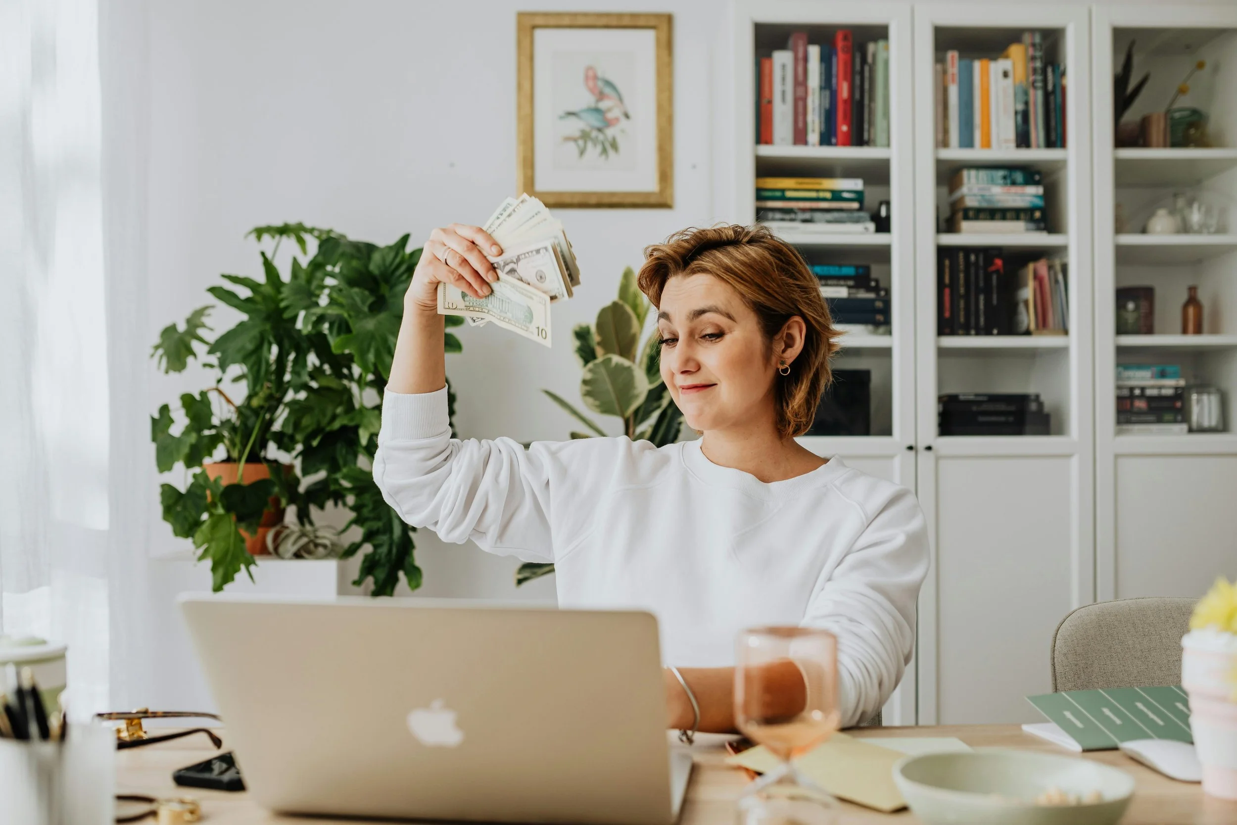 A woman sitting at a desk holding a fan of US dollar bills in her right hand, with a smirk. She has short light brown hair and is wearing a white sweatshirt. The desk has a laptop, a pink glass, and some papers. The background features potted plants, a bookshelf with books, and a framed picture of a bird.