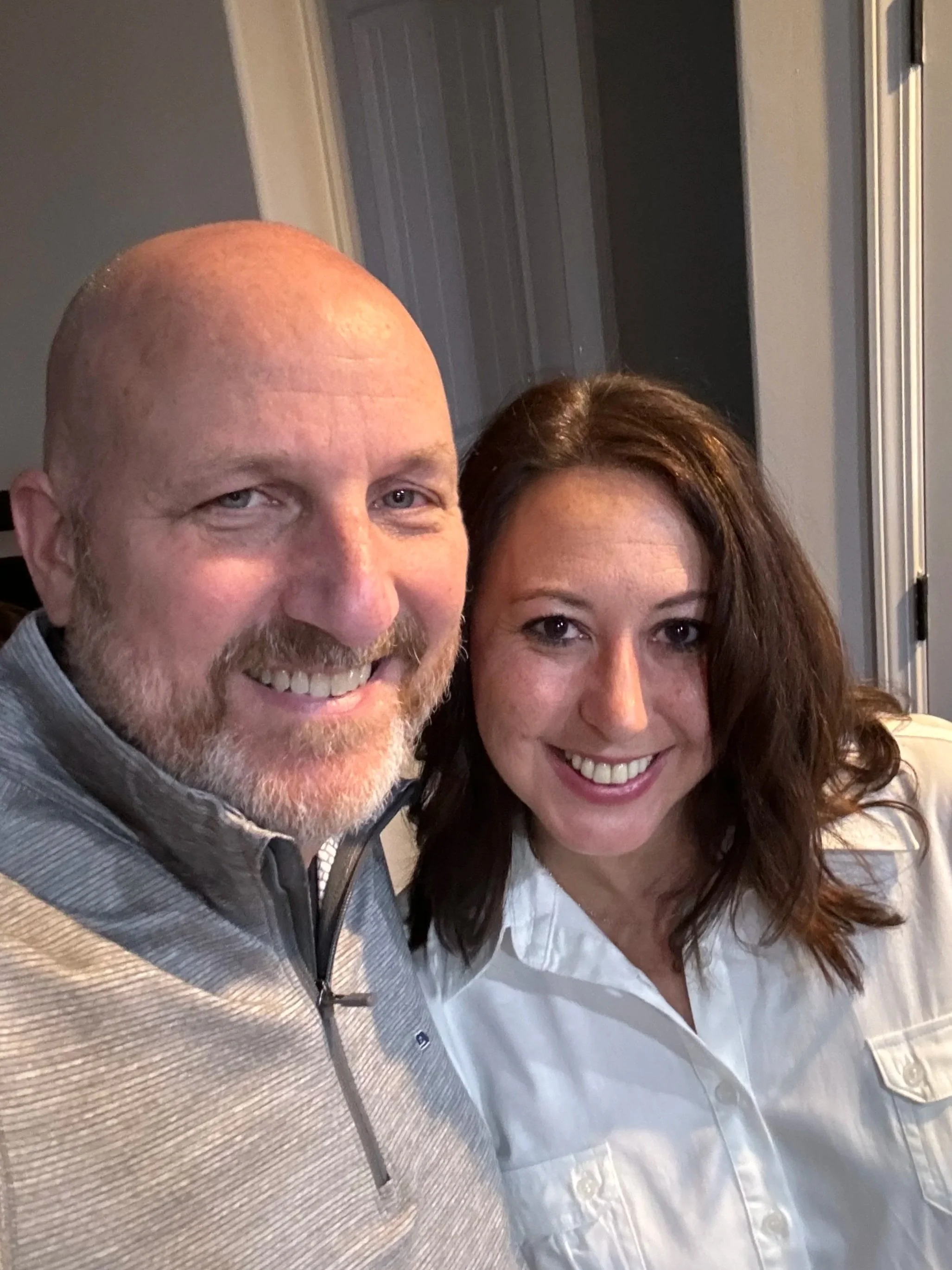 A smiling man with a beard and a woman with brown hair posing together indoors.