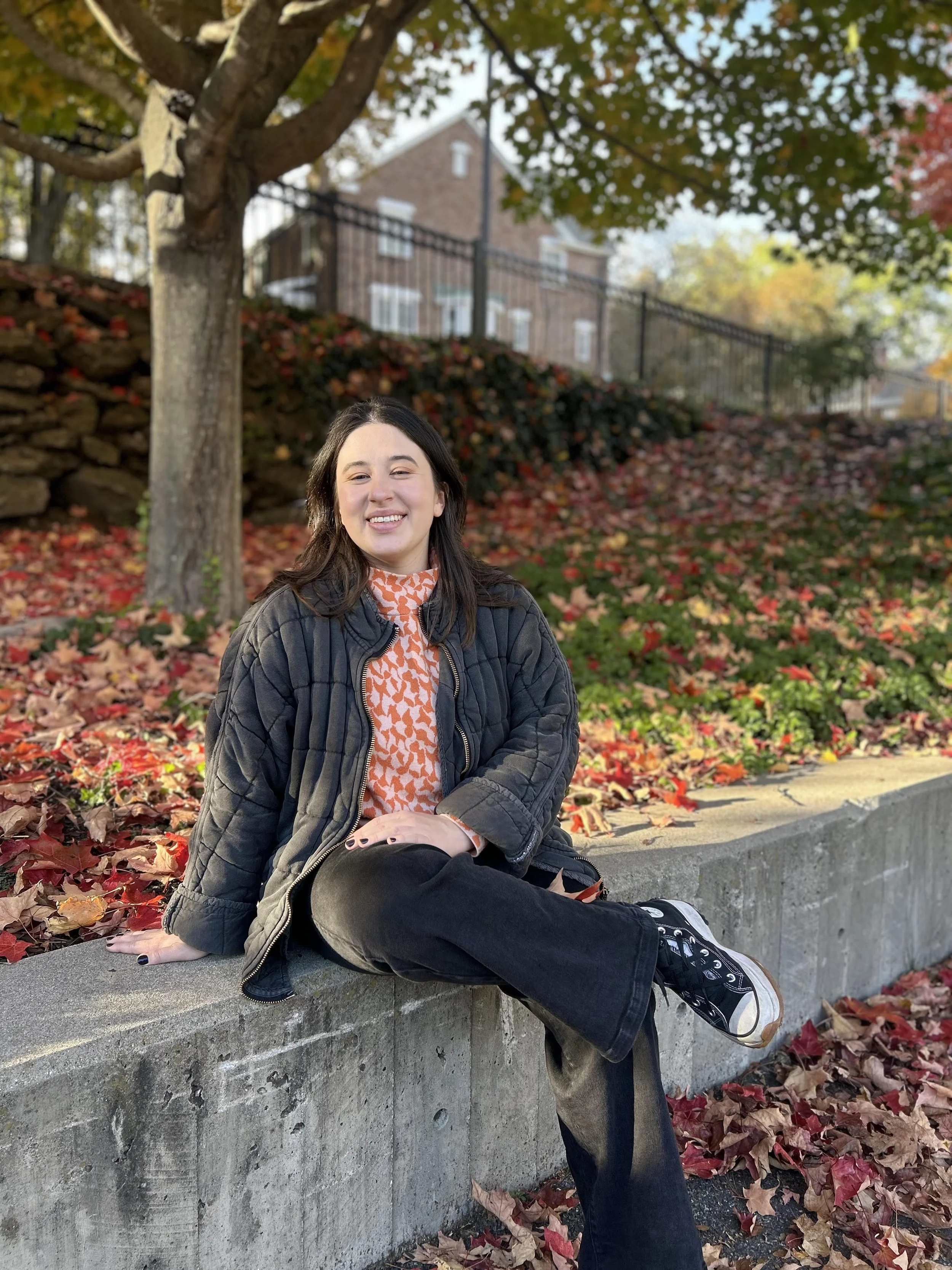 A young woman sitting on a concrete ledge in a park during autumn, surrounded by fallen colorful leaves and waving trees. She is smiling, wearing a black quilted jacket, patterned orange and white shirt, black pants, and black sneakers.