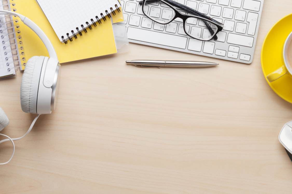 Office desk with headphones, notebooks, glasses, pen, keyboard, yellow coffee mug, and a computer mouse.