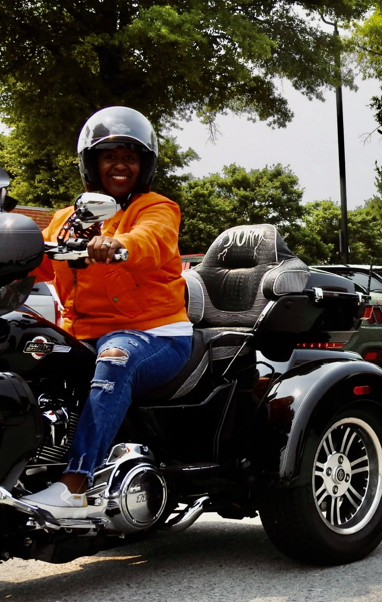 A woman wearing a helmet and orange jacket sitting on a black Harley-Davidson motorcycle with a sidecar, smiling at the camera with trees and cars in the background.