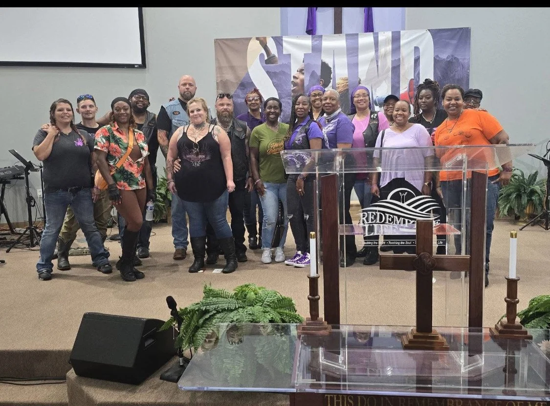Group of diverse people standing together on a church stage with a cross, plants, and a backdrop that says 'STAND' in the background.