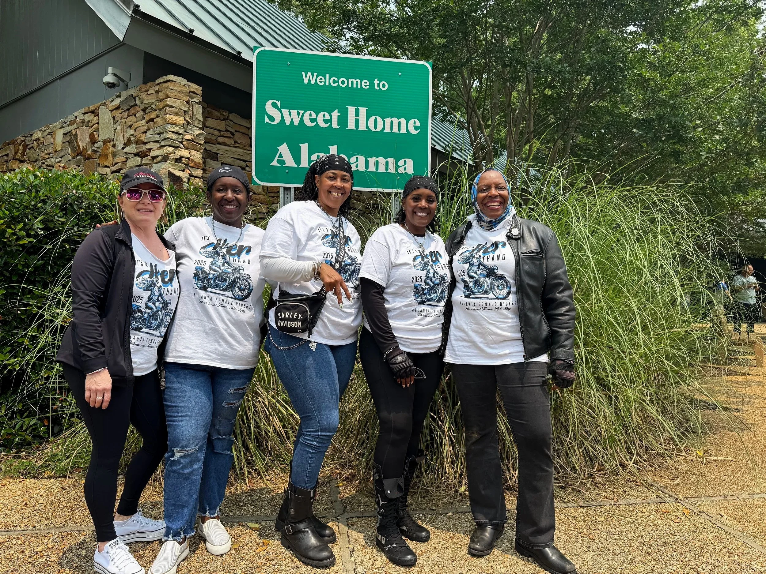 Five women stand together smiling in front of a green