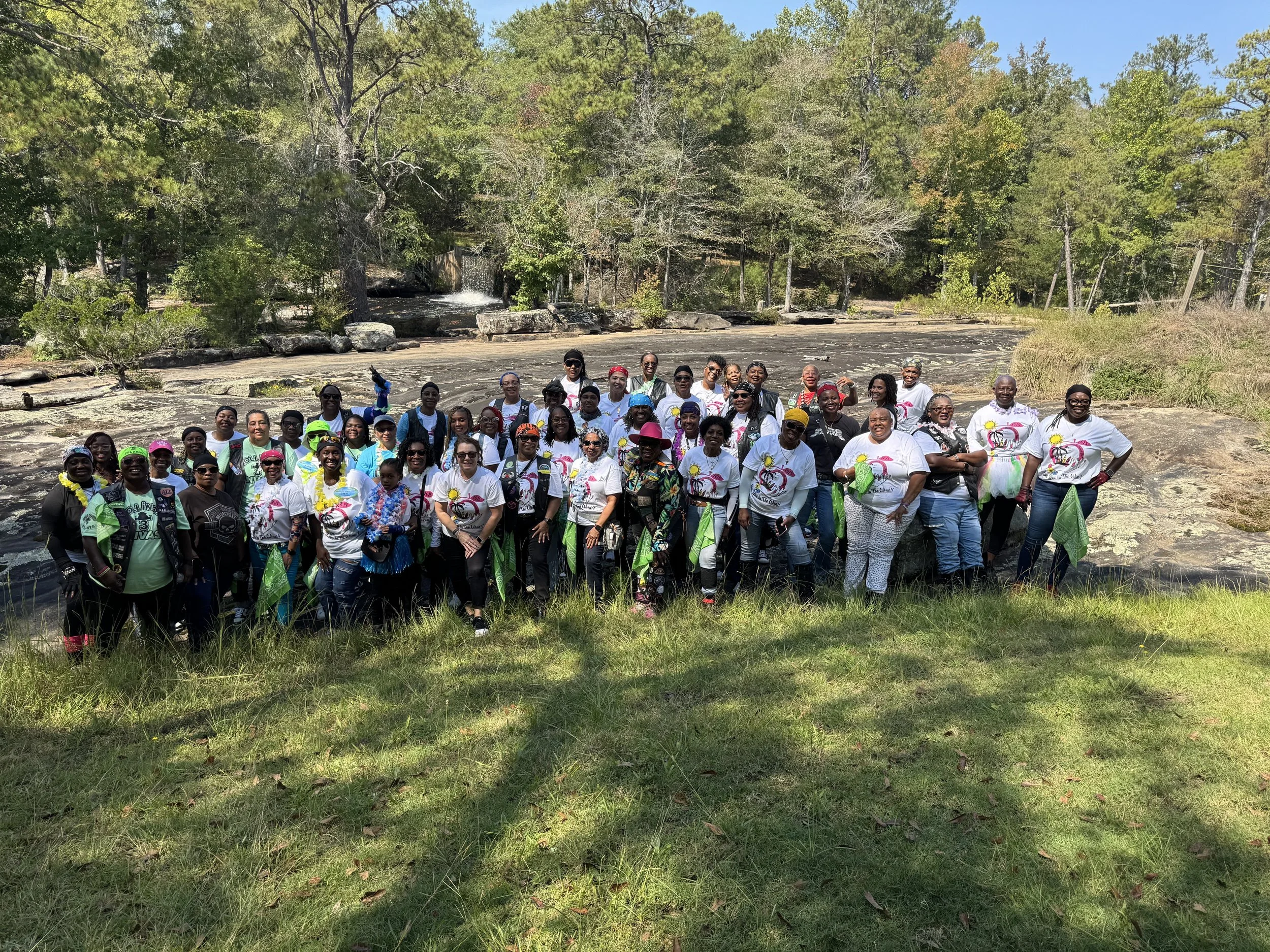 A large group of people posing outdoors near a river or waterfall, with trees and greenery in the background. Many are wearing matching white t-shirts with colorful designs, and some are holding green bandanas or wearing hats.